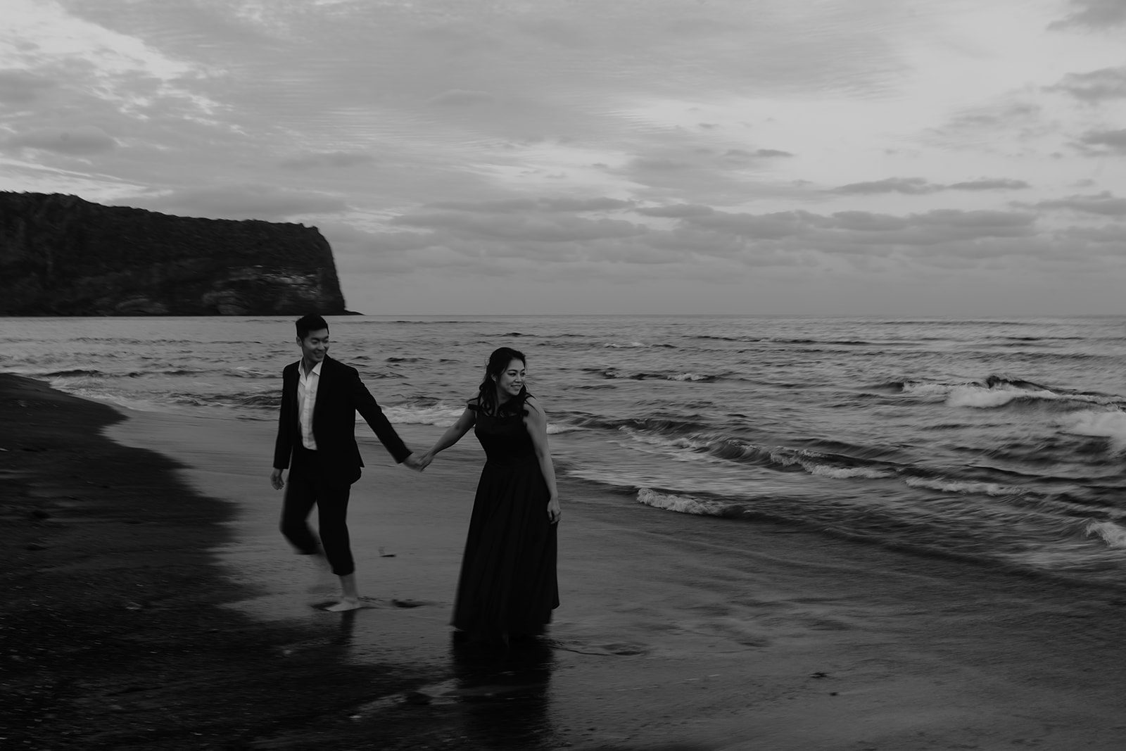 A couple, dressed semi-formally, walks barefoot on a beach along the shoreline at sunset during their Jeju Island pre-wedding shoot. The man wears a suit, and the woman wears a long dress. The scene features waves and a distant rocky cliff.