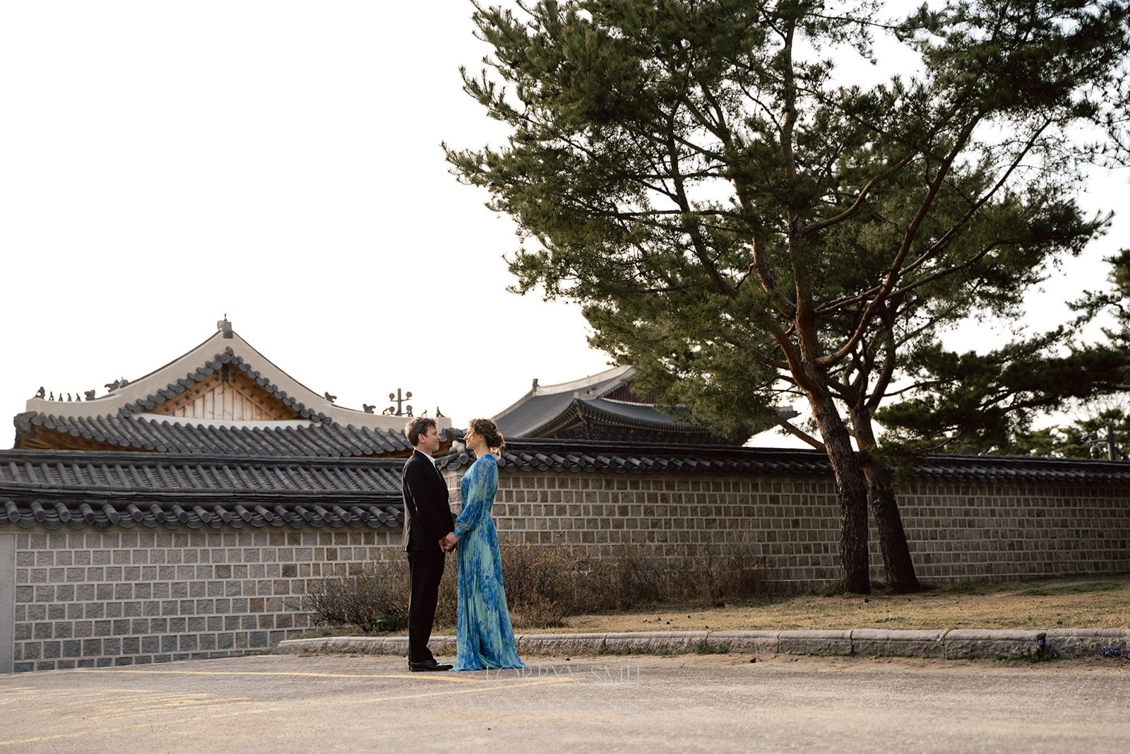 A couple stands holding hands in front of a traditional Korean-style stone wall and tiled rooftops, with a large pine tree nearby.