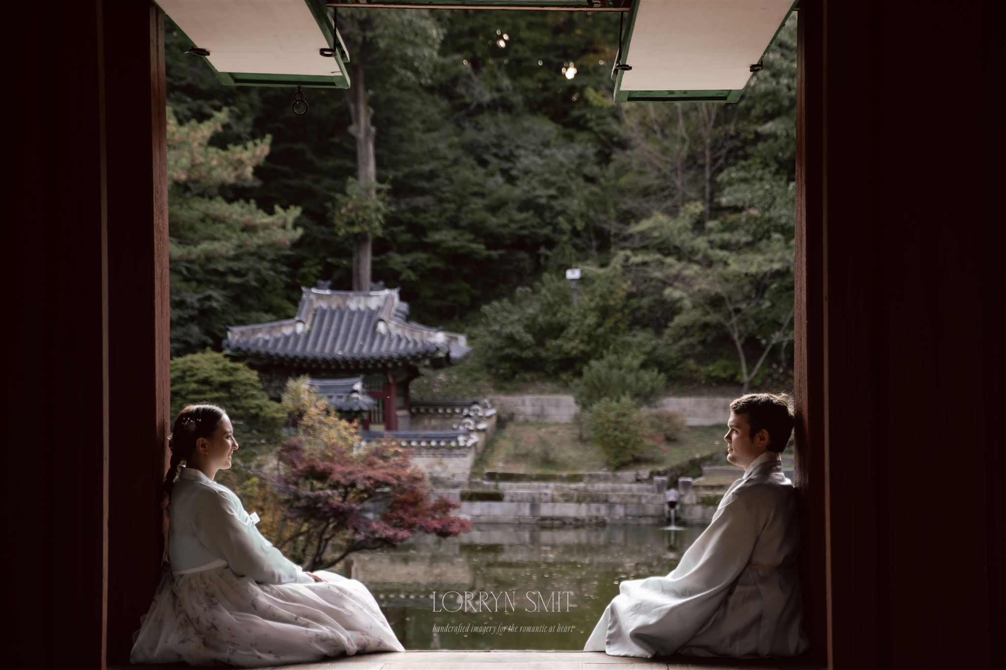 Two people in traditional clothing sit facing each other in a wooden pavilion, overlooking a serene pond and a pagoda—capturing the beauty of how to get married in Korea amid lush greenery.