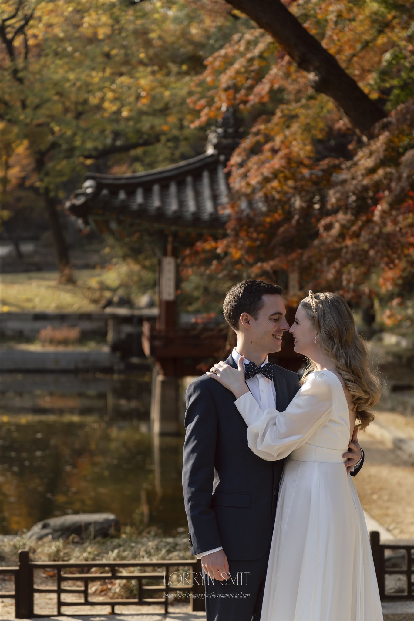 A couple dressed formally embrace and smile at each other in front of a traditional pavilion, with autumn foliage and a pond in the background.