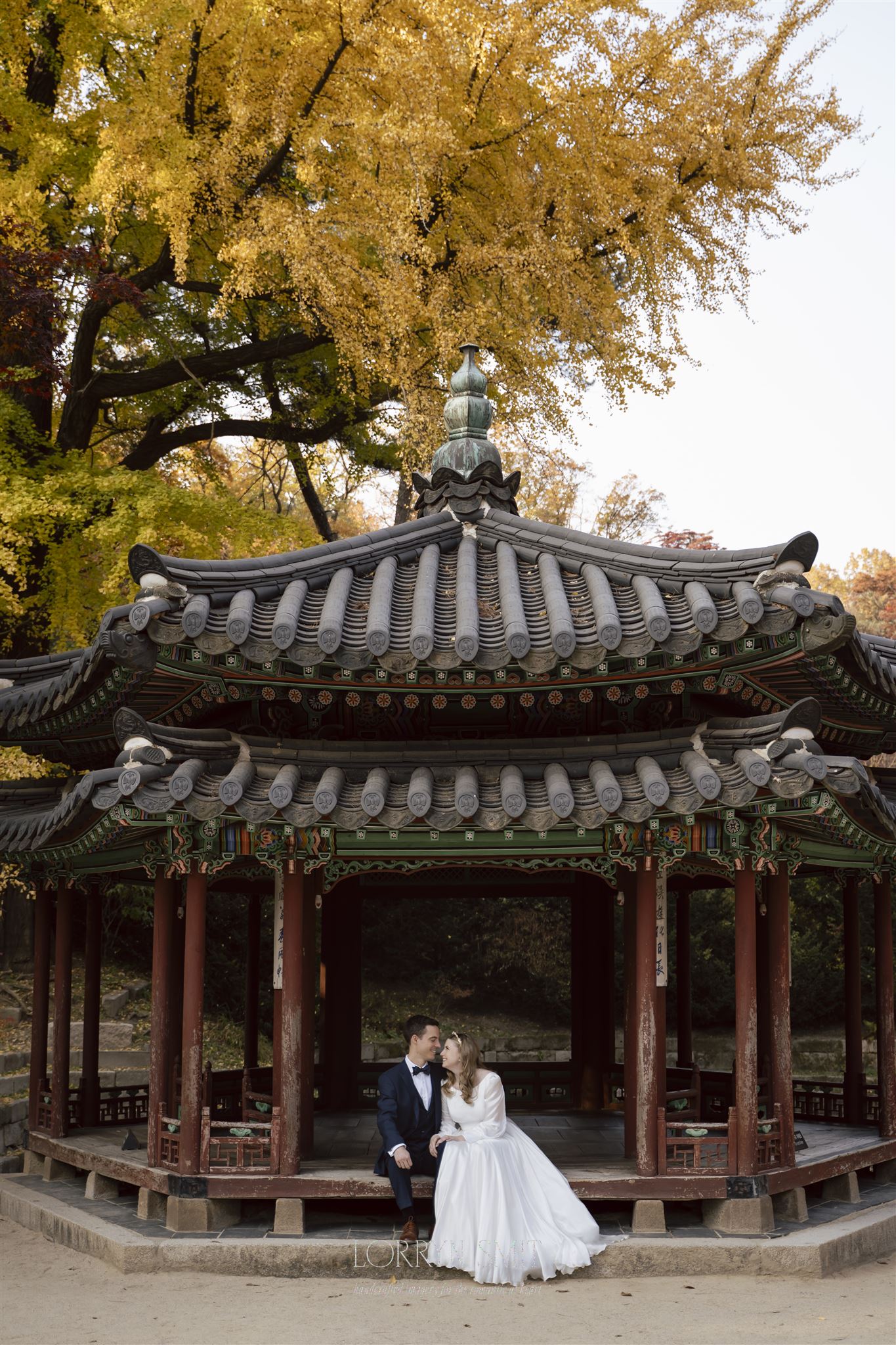 A bride and groom sit together under a traditional Korean pavilion with autumn trees in the background, offering a picturesque glimpse into how to get married in Korea.