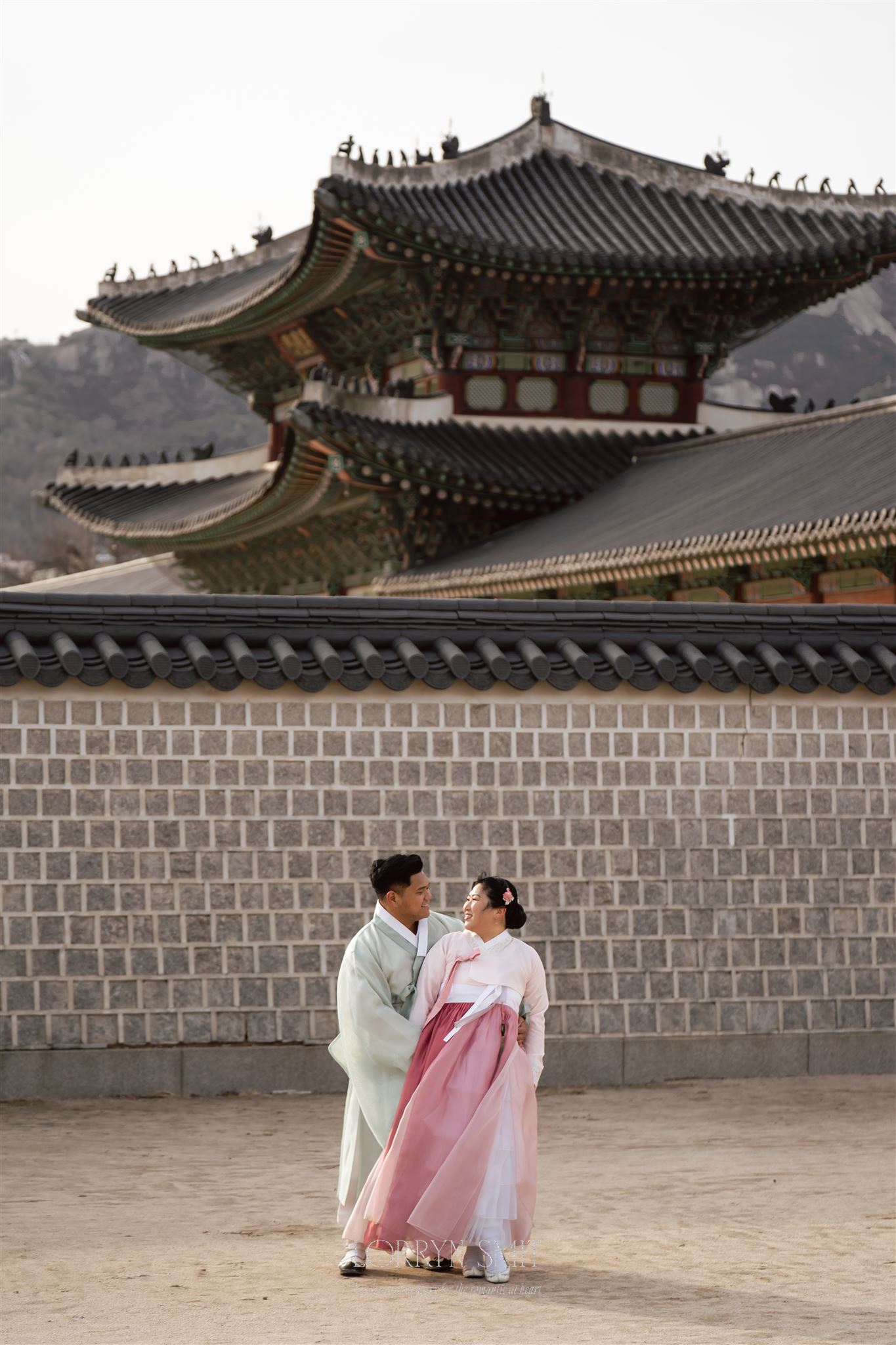 Two people in traditional Korean hanbok stand in front of a stone wall with a historic palace building in the background, evoking the elegance of ceremonies for those interested in how to get married in Korea.