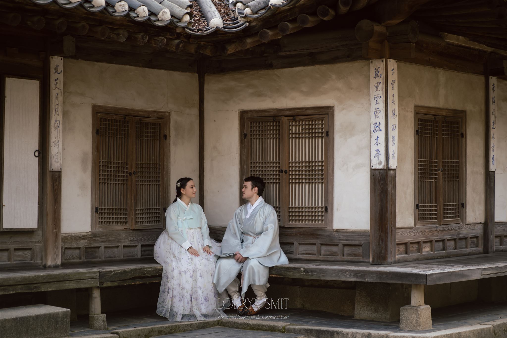 A man and a woman in traditional Korean hanbok sit on a wooden bench outside a historic building with wooden doors and tile roof, evoking the elegance of those learning how to get married in Korea.