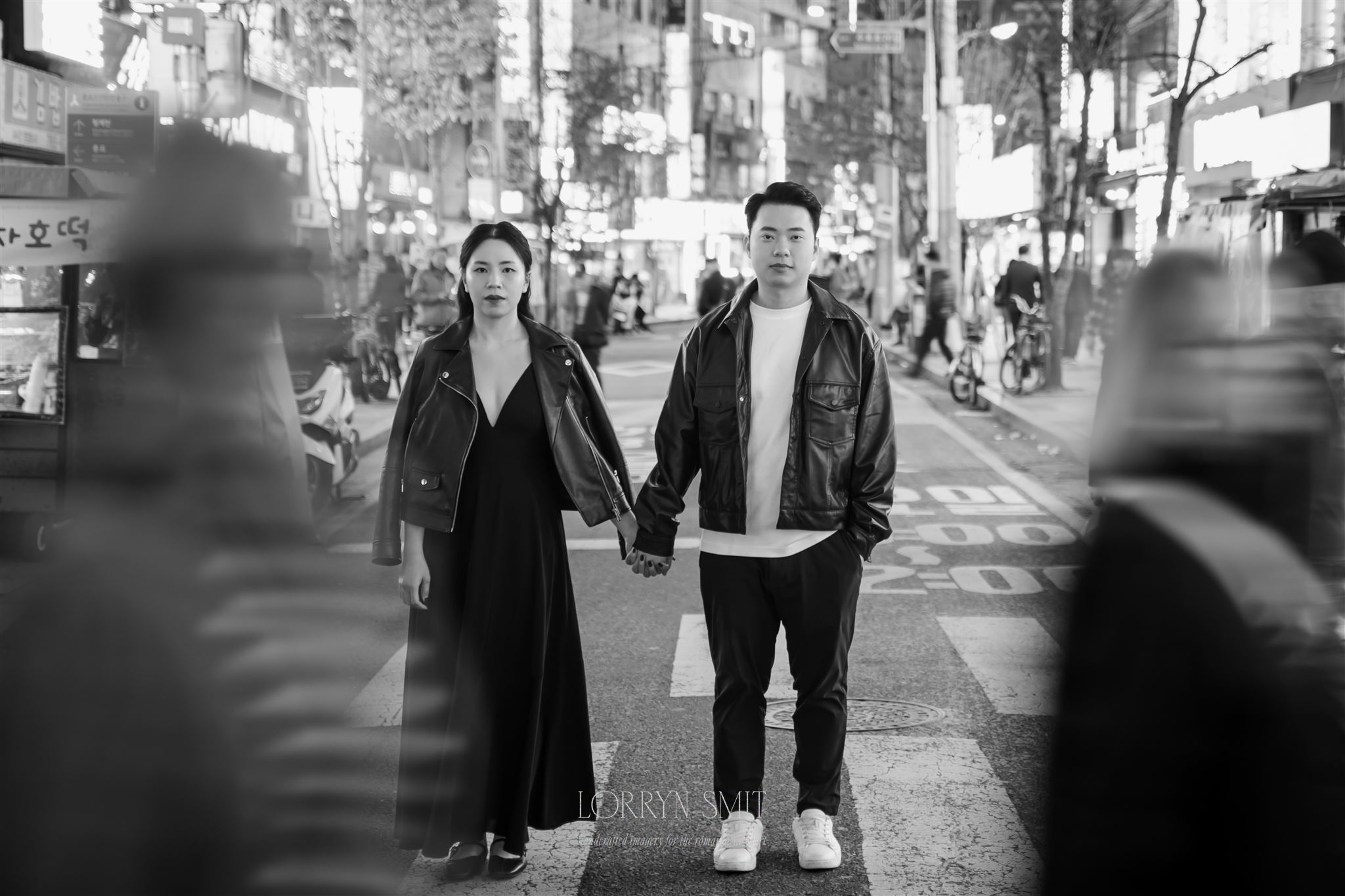 A man and woman holding hands stand still on a busy city street at night as blurred pedestrians pass by, after they get married in Korea amid the city lights.