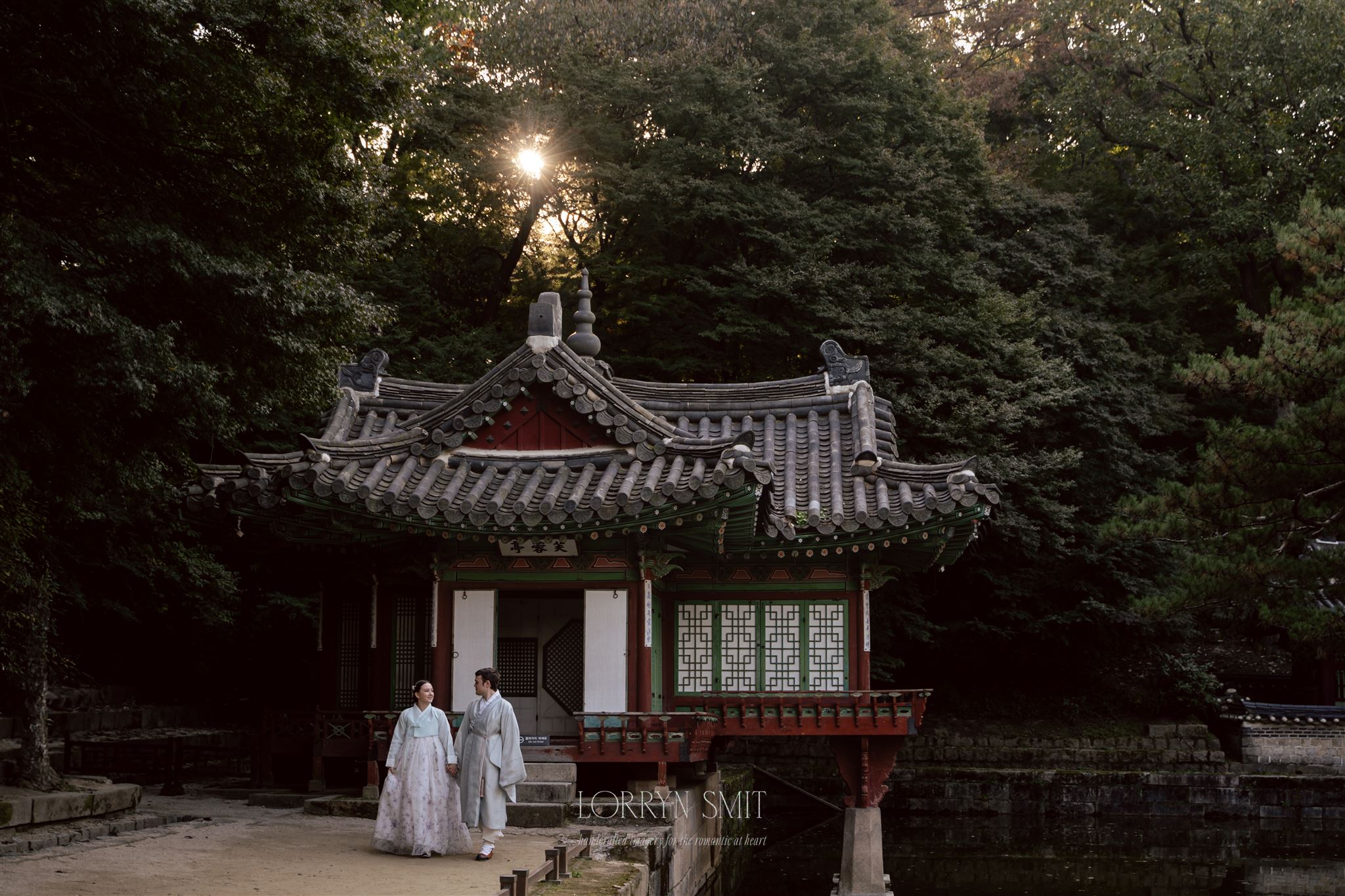 Two people in traditional Korean clothing walk near a historic wooden pavilion, surrounded by lush trees and soft sunlight, a picturesque scene reminiscent of how to get married in Korea through time-honored customs.