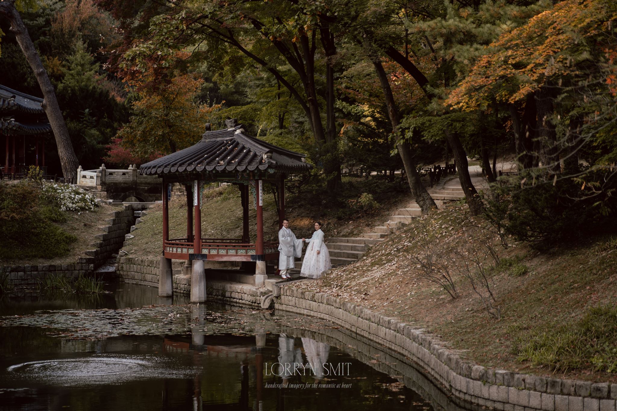 Two people in traditional Korean attire stand by a small pavilion next to a pond, surrounded by autumn foliage, reflecting the beauty of how to get married in Korea amidst nature’s splendor.