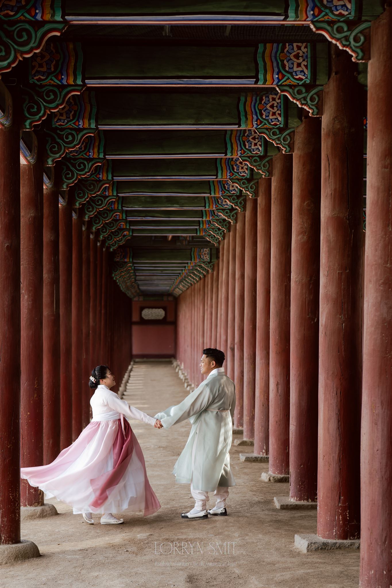 A couple dressed in traditional Korean hanbok holds hands and dances in a corridor with red columns and ornate painted beams, capturing a timeless moment that reflects the beauty of getting married in South Korea.