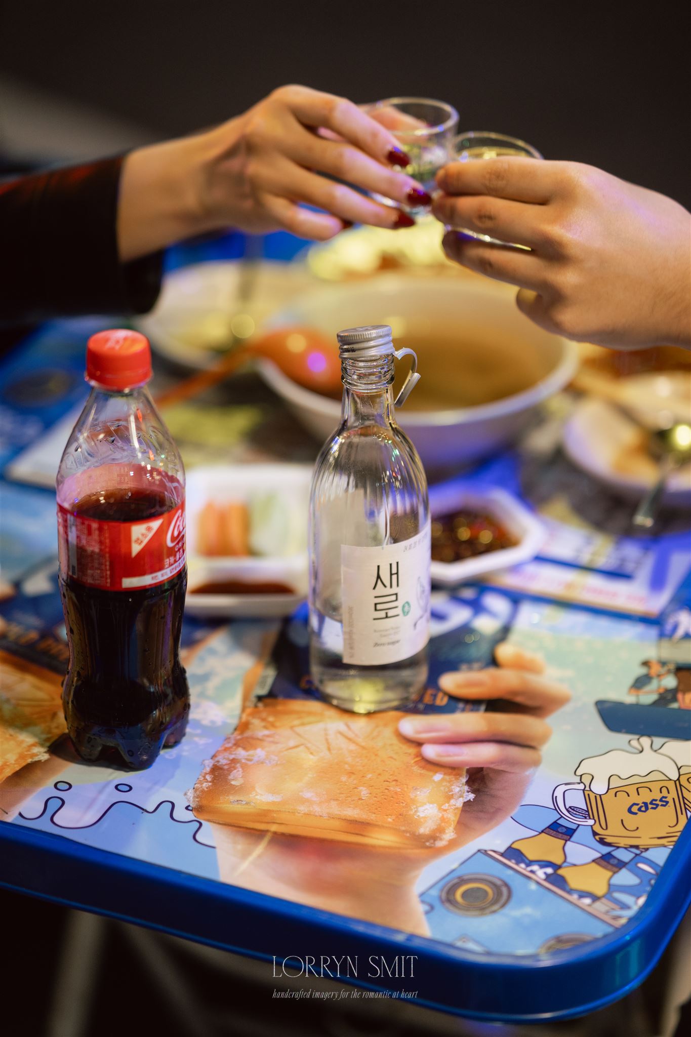 Two people toast with small glasses over a table featuring soju, Coca-Cola, food dishes, and an illustrated placemat, celebrating their elopement in Korea.