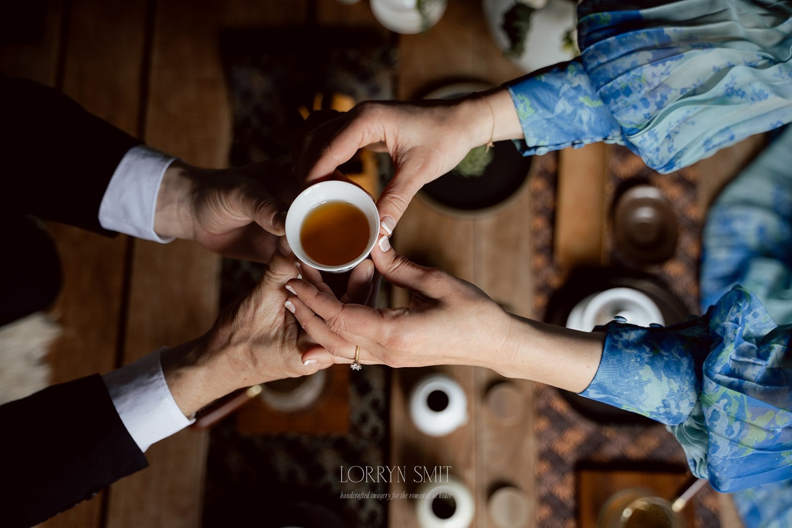 Two people hold a small cup of tea together over a wooden table set with teaware, seen from above—a moment reminiscent of a Korean wedding tea ceremony.