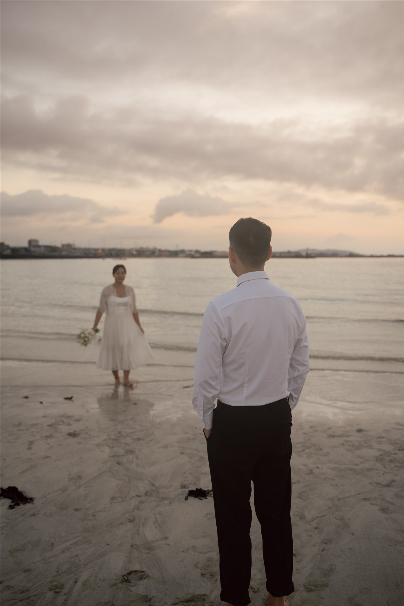 A man in a white shirt and black pants stands on a beach, facing a woman in a white dress holding flowers during their pre wedding photoshoot in Korea, with calm water and a cloudy sky in the background.