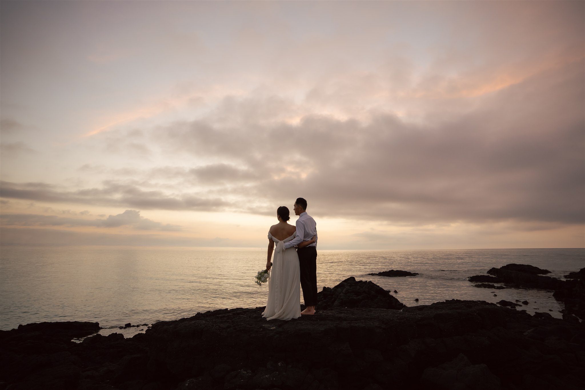 A couple in formal attire stands on a rocky shore at sunset under a cloudy sky—a timeless moment from their pre wedding photoshoot in Korea.