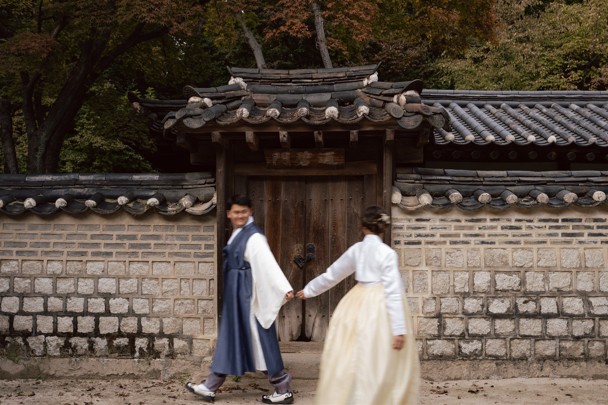 Two people in traditional Korean hanbok walk hand in hand past a wooden gate and stone wall, with trees and foliage in the background—an elegant scene from a pre wedding photoshoot in Korea.