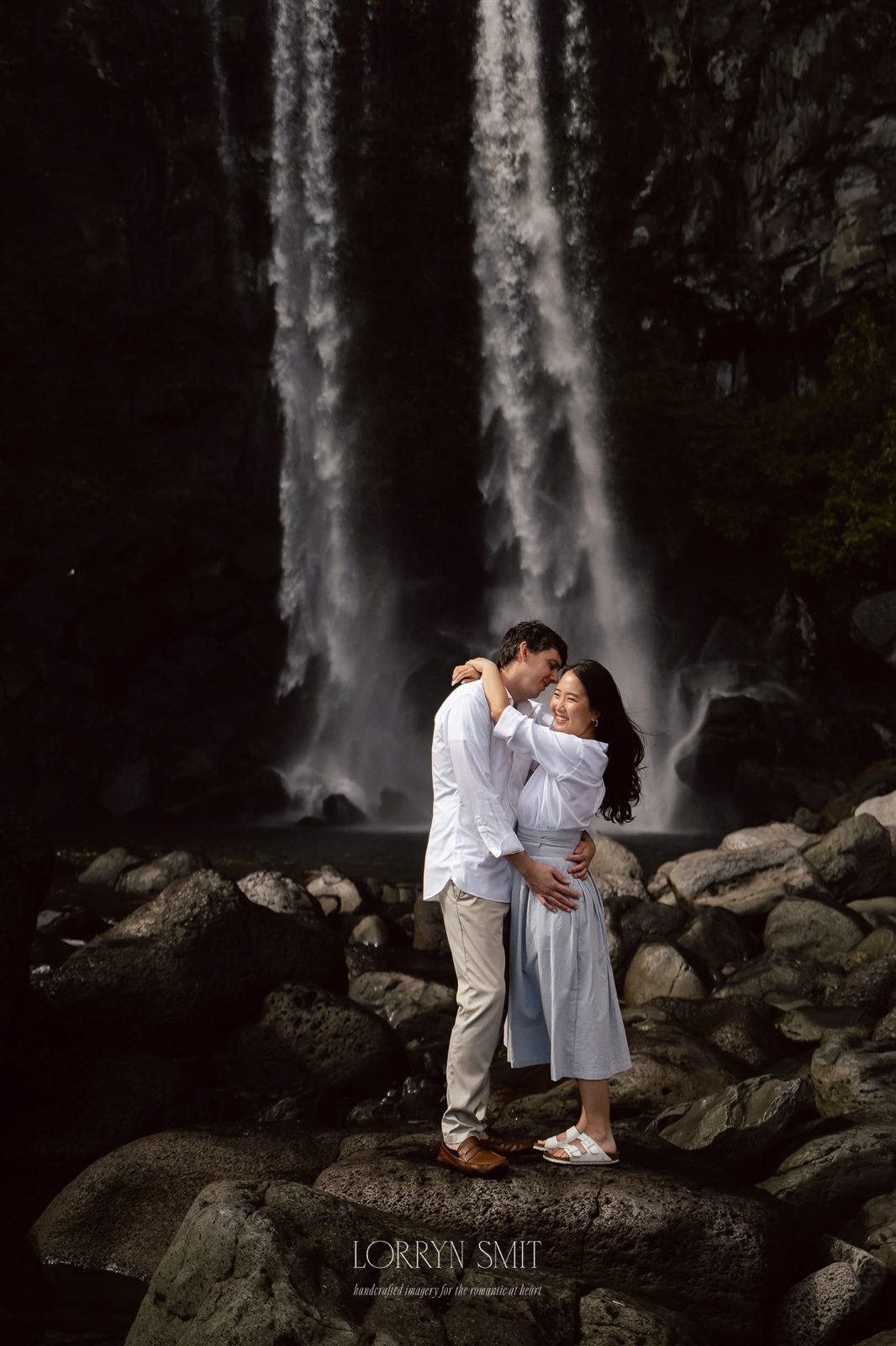 A couple embraces on rocky ground in front of a tall, cascading waterfall during their pre wedding photoshoot in Korea.