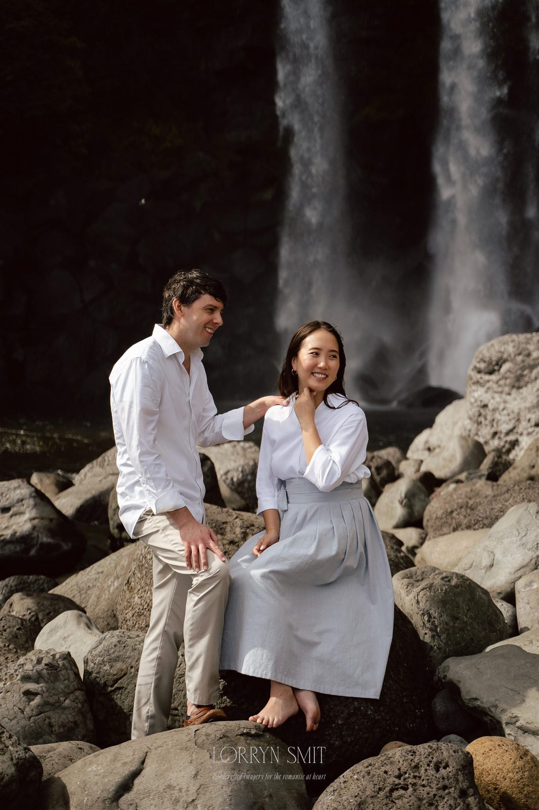 A man and woman sit together on rocks near a waterfall, both smiling in light-colored clothing during their pre wedding photoshoot in Korea.