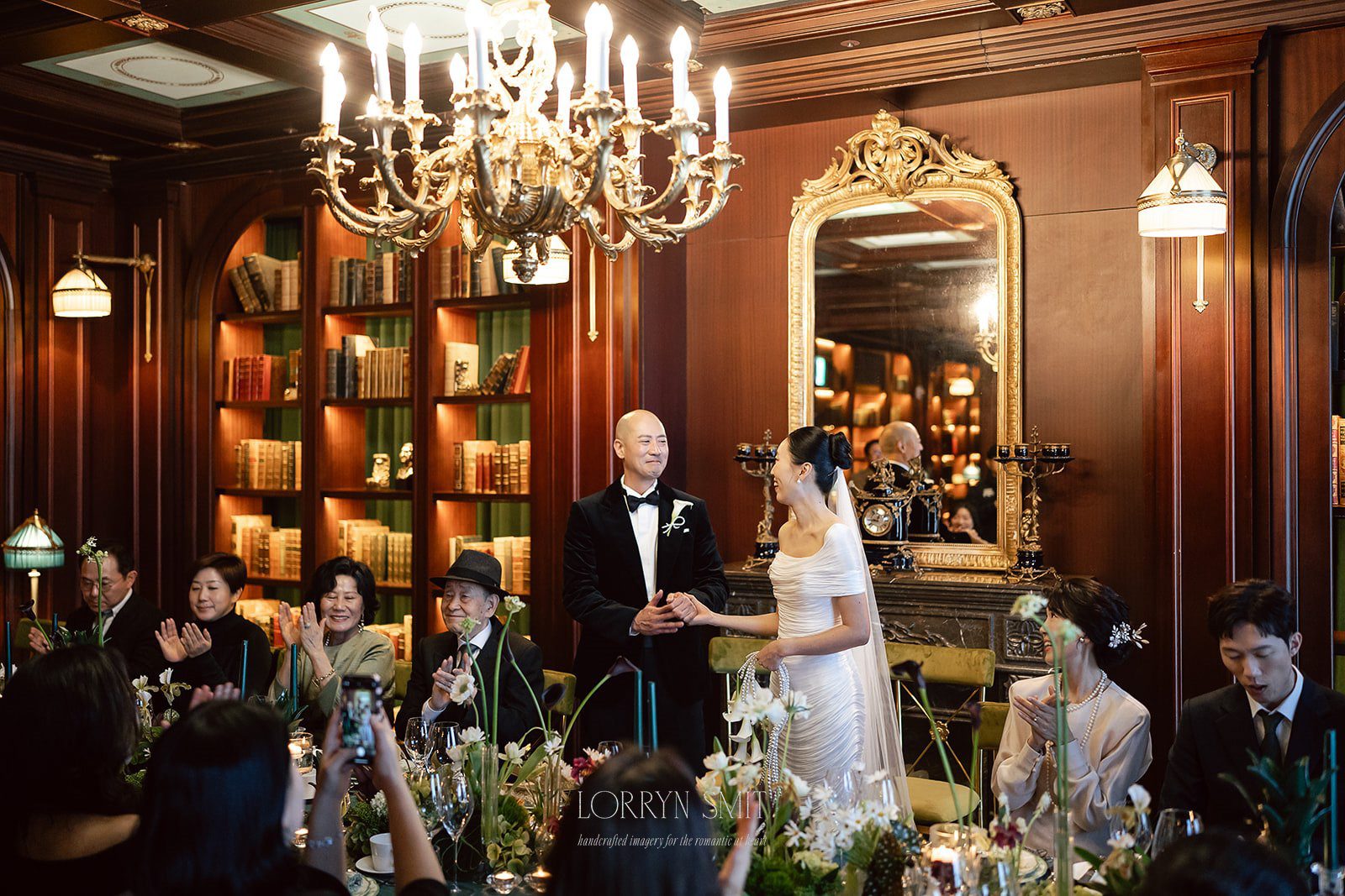 A bride and groom stand together in an elegant, wood-paneled room at L’Escape Hotel, surrounded by seated guests who are clapping and smiling during a micro wedding reception.