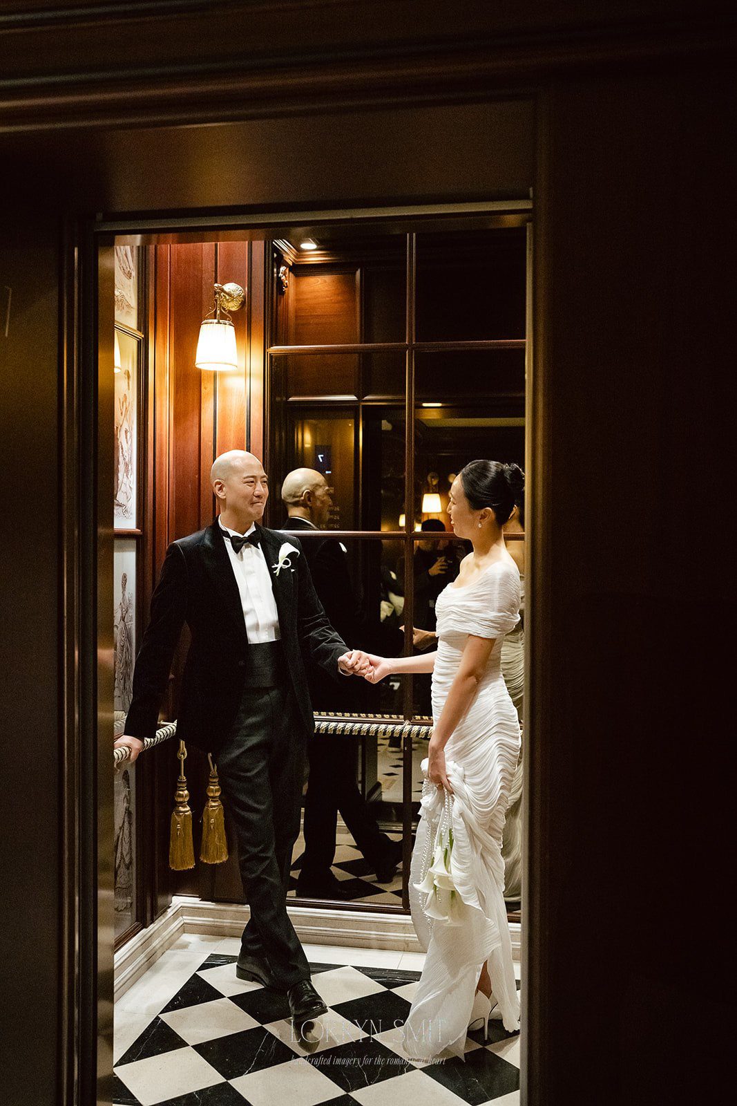 A couple in formal attire stands holding hands in an elegant, mirrored elevator with black and white checkered flooring, capturing a stylish moment from their micro wedding at L'Escape Hotel.