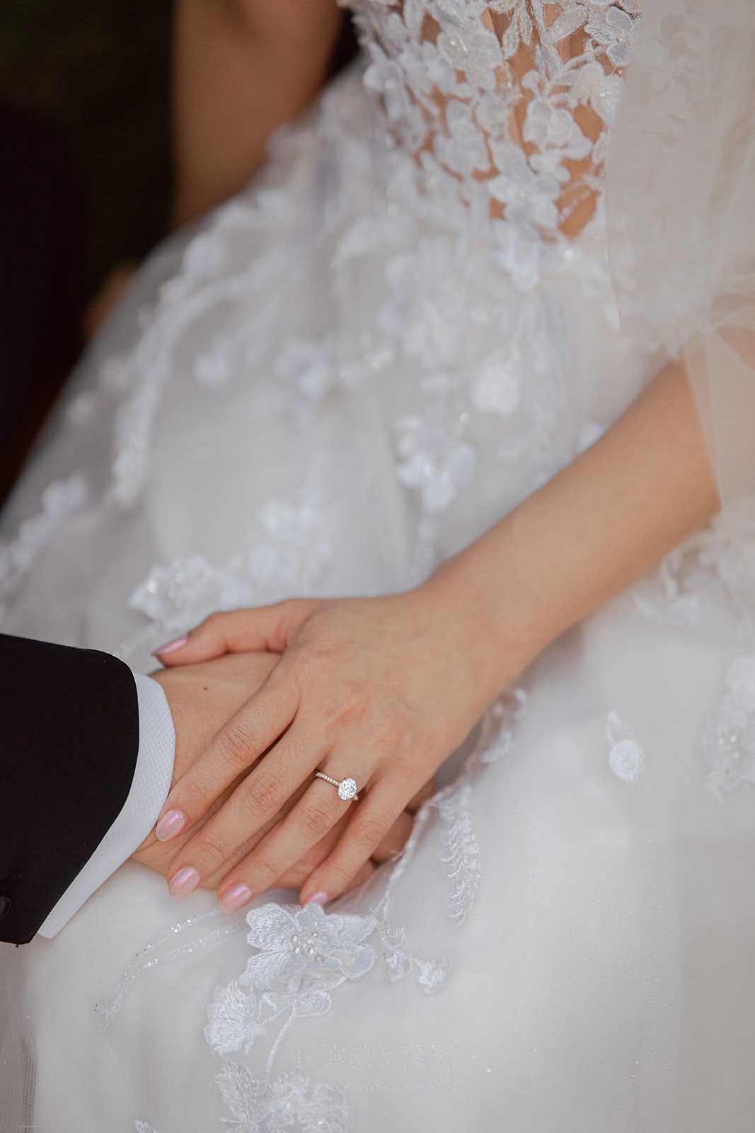Close-up of a bride’s hand with an engagement ring resting on a groom’s hand, both in formal attire with lace wedding dress details.