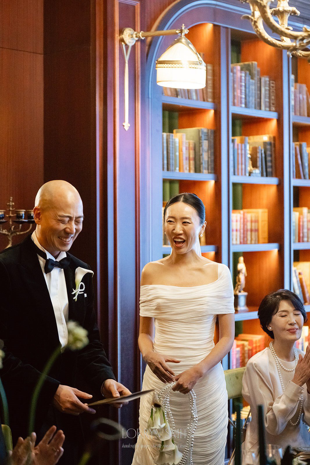 A bride and groom stand smiling together indoors, with bookshelves and seated guests in the background—capturing a scene a wedding content creator might highlight to showcase personal moments from the big day.
