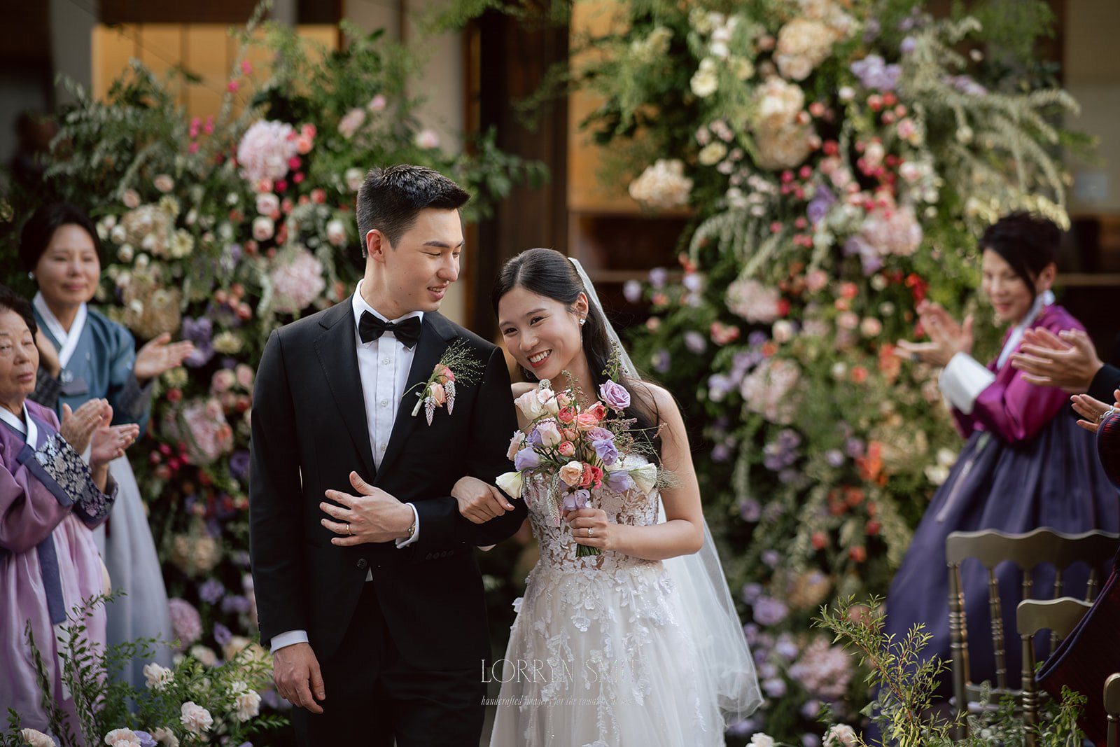 A bride and groom walk arm in arm, smiling, surrounded by floral arrangements and guests clapping—moments a wedding content creator would capture, with some attendees wearing traditional Korean attire.