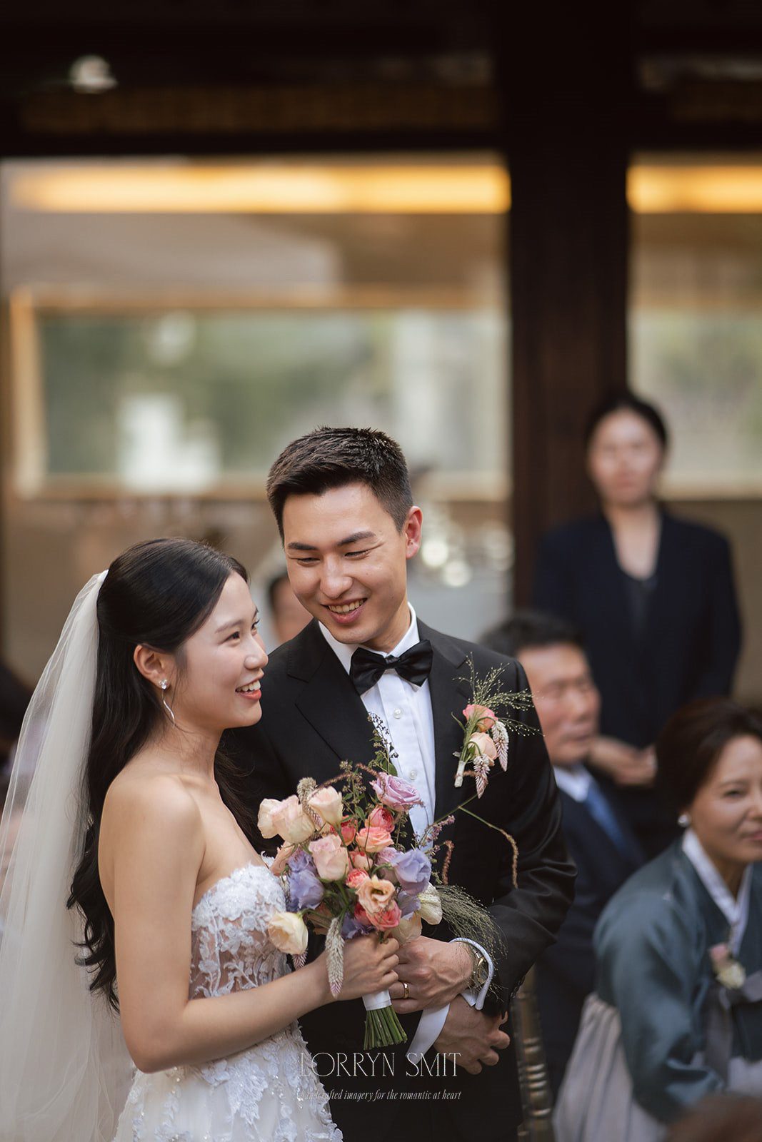 A bride and groom in formal attire smile at each other while holding a bouquet at an outdoor wedding ceremony, with guests seated in the background.