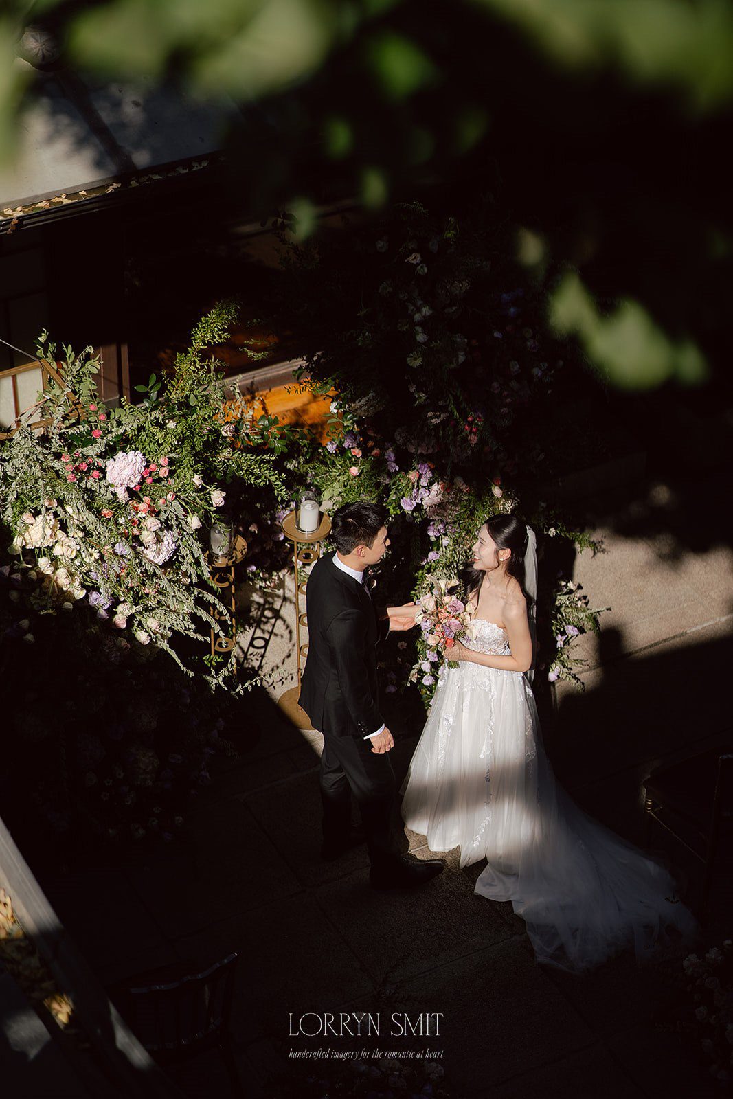 A bride and groom stand facing each other outdoors, surrounded by floral arrangements and dappled sunlight, while a wedding content creator captures their special moment.