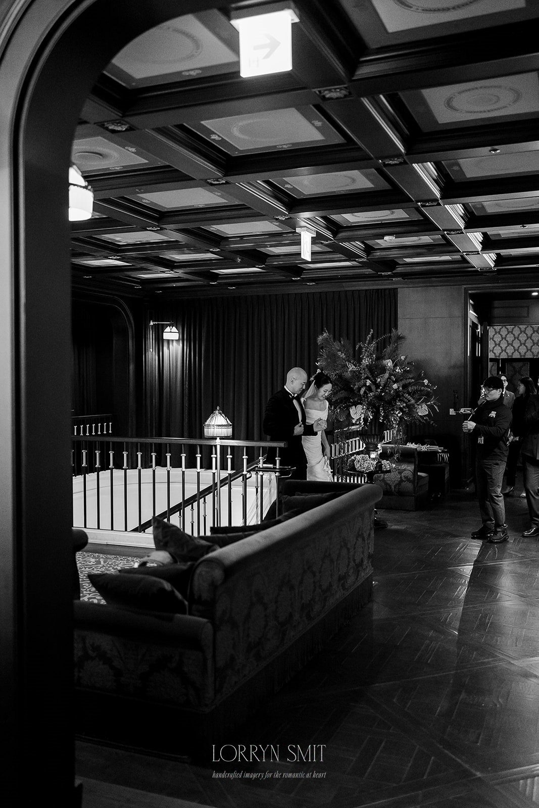A couple stands together near a railing in an elegant, dimly-lit room with patterned ceiling, wooden floor, and plush seating—perhaps capturing moments for a wedding content creator as other guests converse nearby.