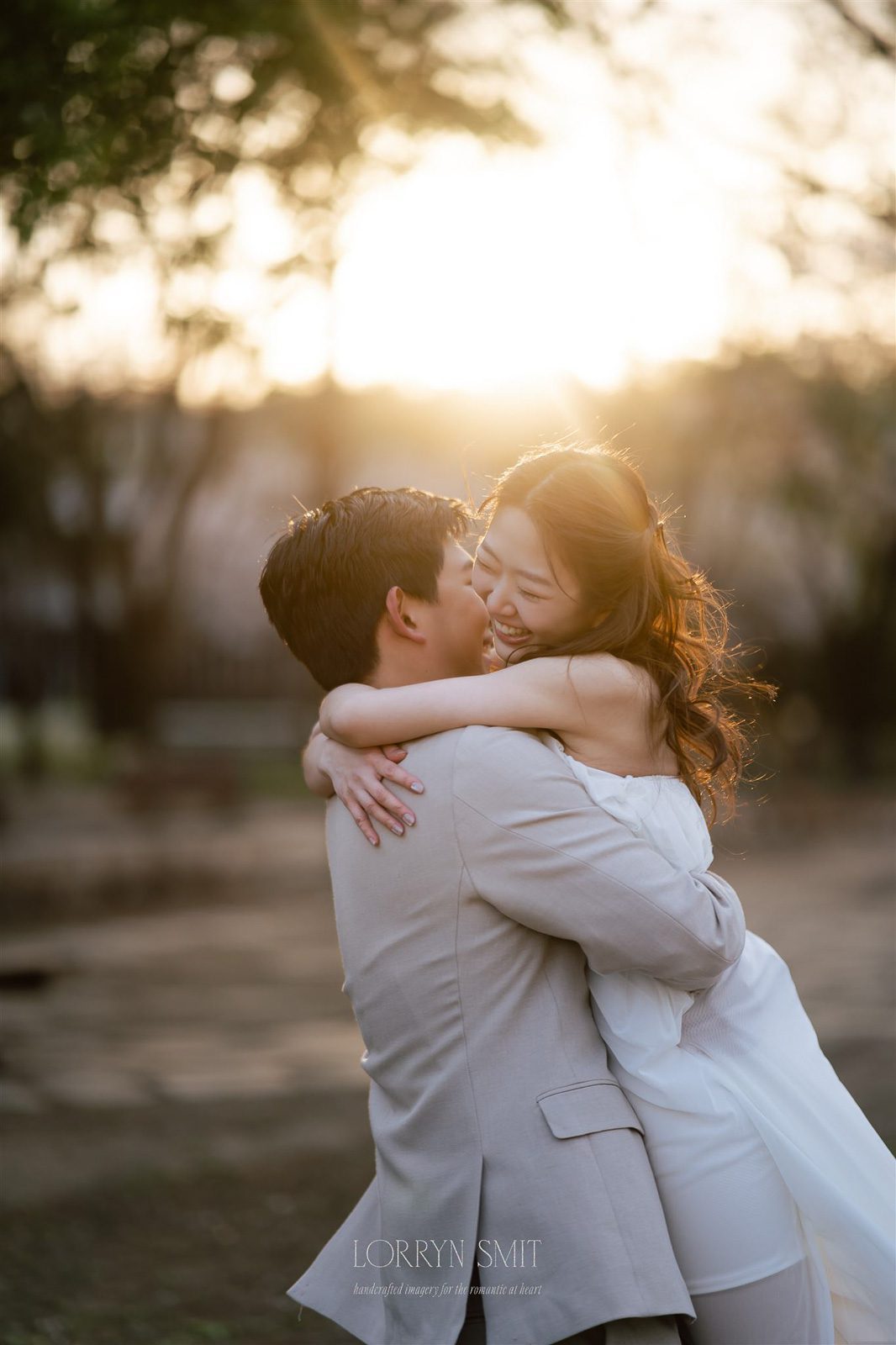 A man in a light suit lifts and hugs a smiling woman in a white dress outdoors at sunset, capturing the joy of their pre wedding photoshoot in Korea.