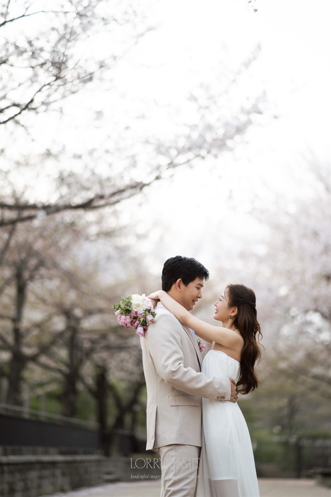 A couple dressed in light-colored formal attire embraces outdoors under blooming cherry blossom trees.