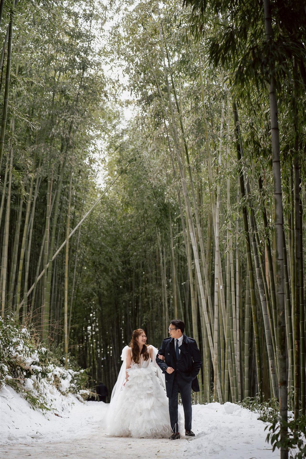 A bride and groom, dressed in wedding attire, walk arm in arm through a snow-covered bamboo forest.