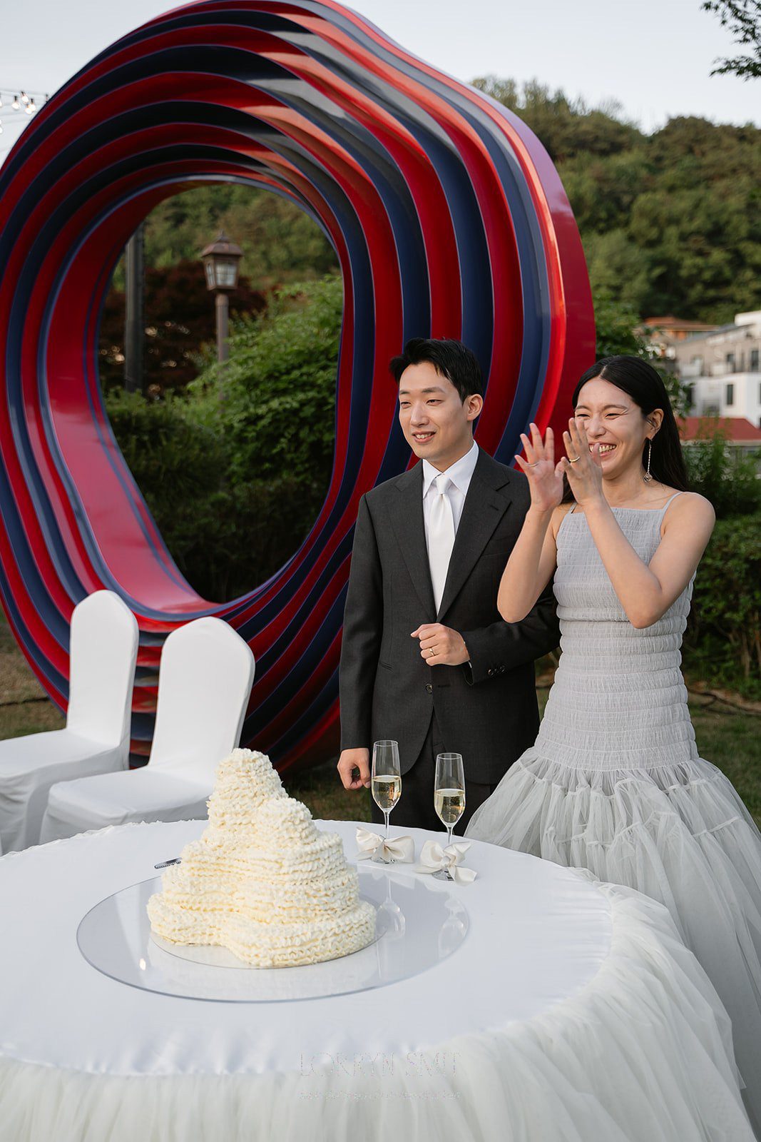 A couple in formal attire stands by a table with a uniquely shaped cake and two glasses of champagne, set against a large abstract sculpture