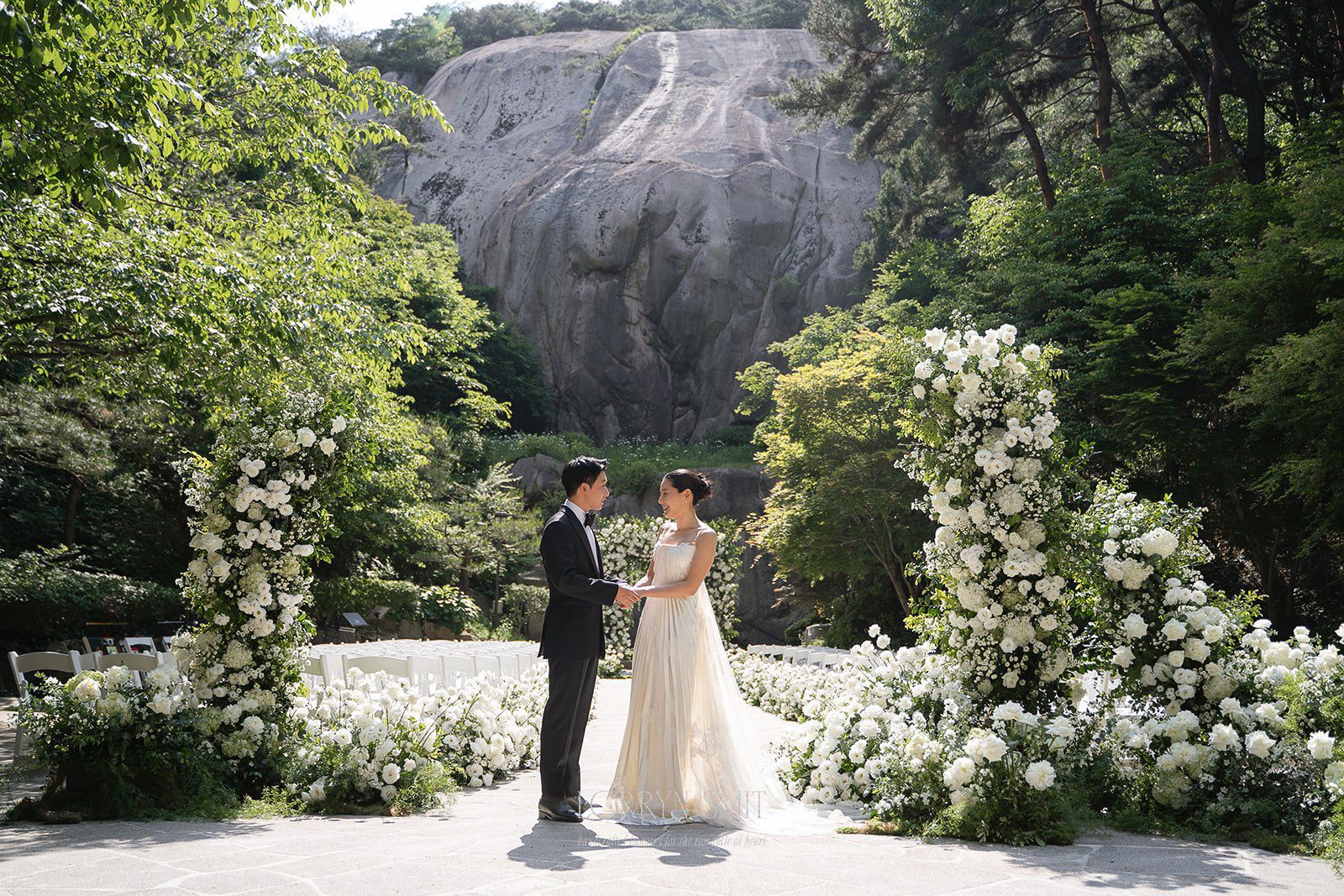 A bride and groom stand facing each other outdoors at one of the stunning wedding venues in Korea, surrounded by white flowers and greenery, with a rocky cliff in the background.