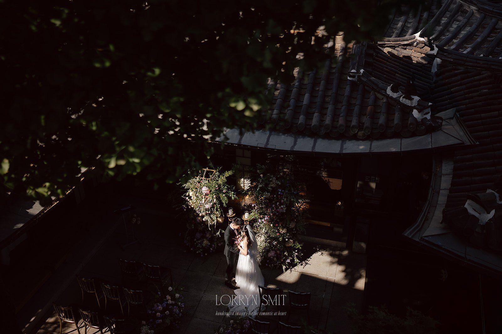 Bride and groom stand together in sunlight near a floral arch in a courtyard, surrounded by tiled rooftops and shadowed tree branches—capturing the romantic charm of wedding venues in Korea.