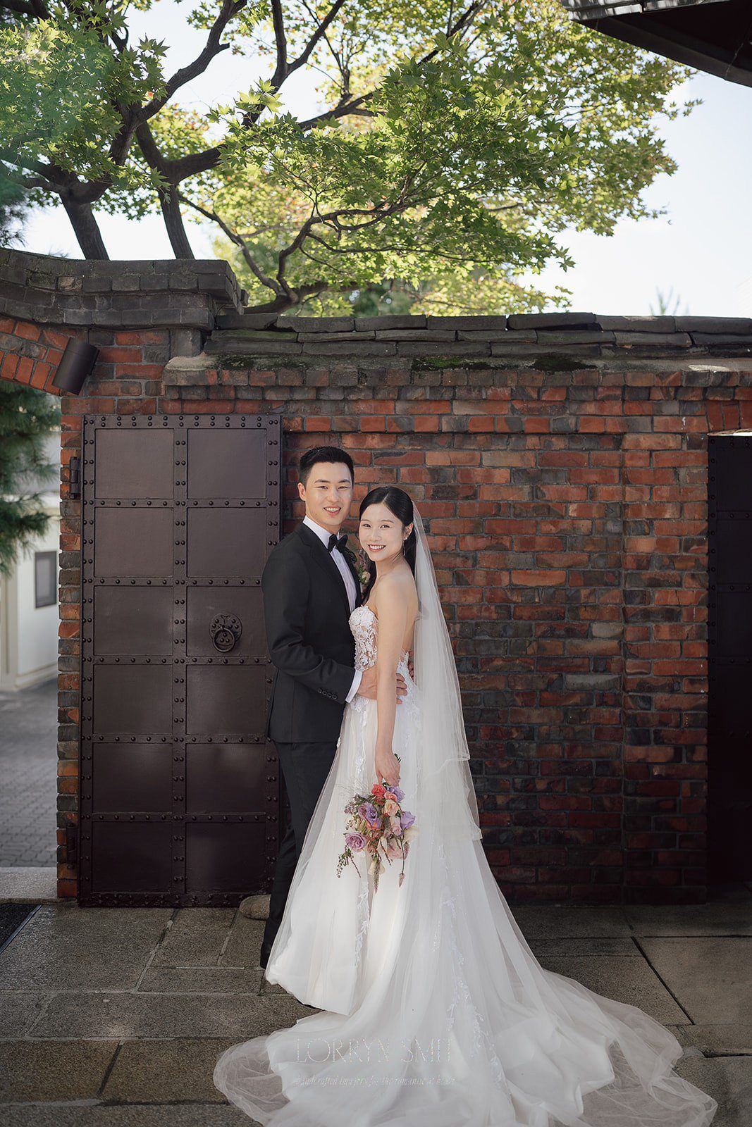 A bride and groom in formal wedding attire stand side by side, smiling, in front of a brick wall and metal gate with trees in the background—capturing the charm often found at wedding venues in Korea.
