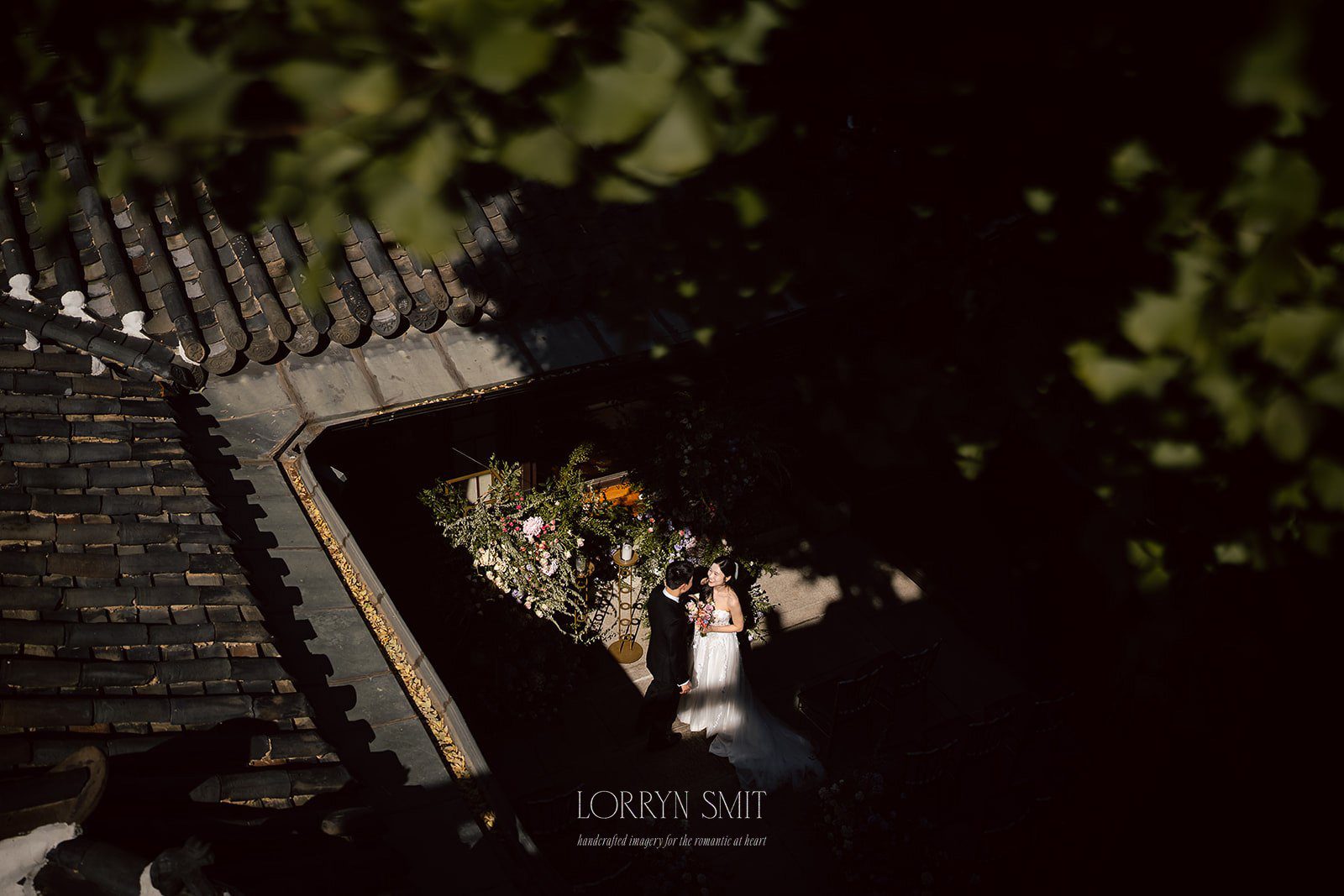 A bride and groom stand together in a sunlit courtyard, surrounded by greenery and flowers, viewed from above—a picture-perfect moment for a Korea destination wedding.