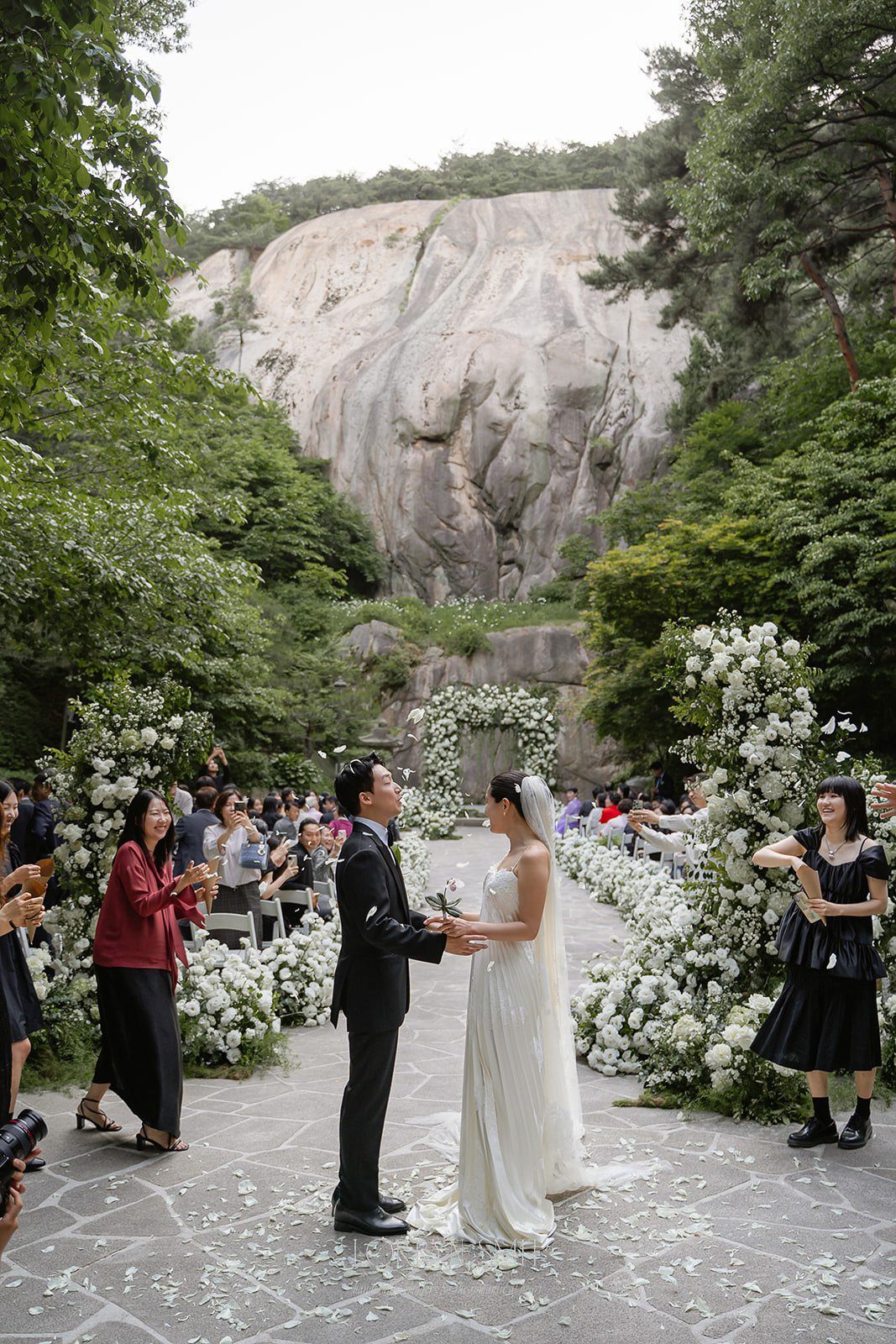 A bride and groom stand facing each other at an outdoor wedding ceremony, surrounded by white flowers and guests, with a rocky cliff and greenery—showcasing one of the most breathtaking wedding venues in Korea.