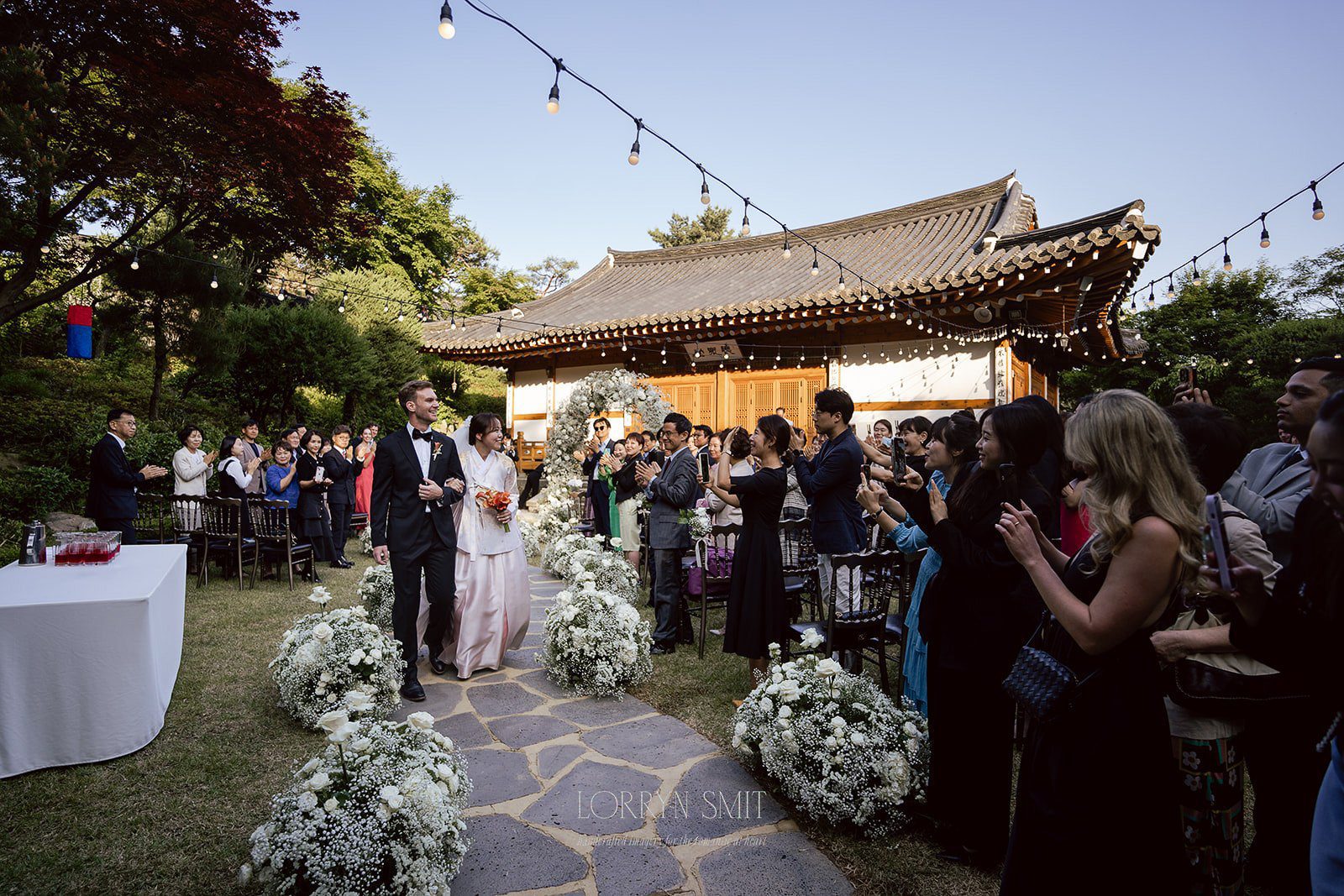 A bride and groom walk down an outdoor aisle lined with flowers, surrounded by guests, at a traditional-style venue decorated with string lights—one of the most picturesque wedding venues in Korea.
