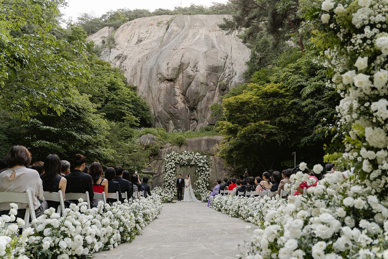 A wedding ceremony with guests seated on either side of a flower-lined aisle, set against a backdrop of trees and a large boulder.