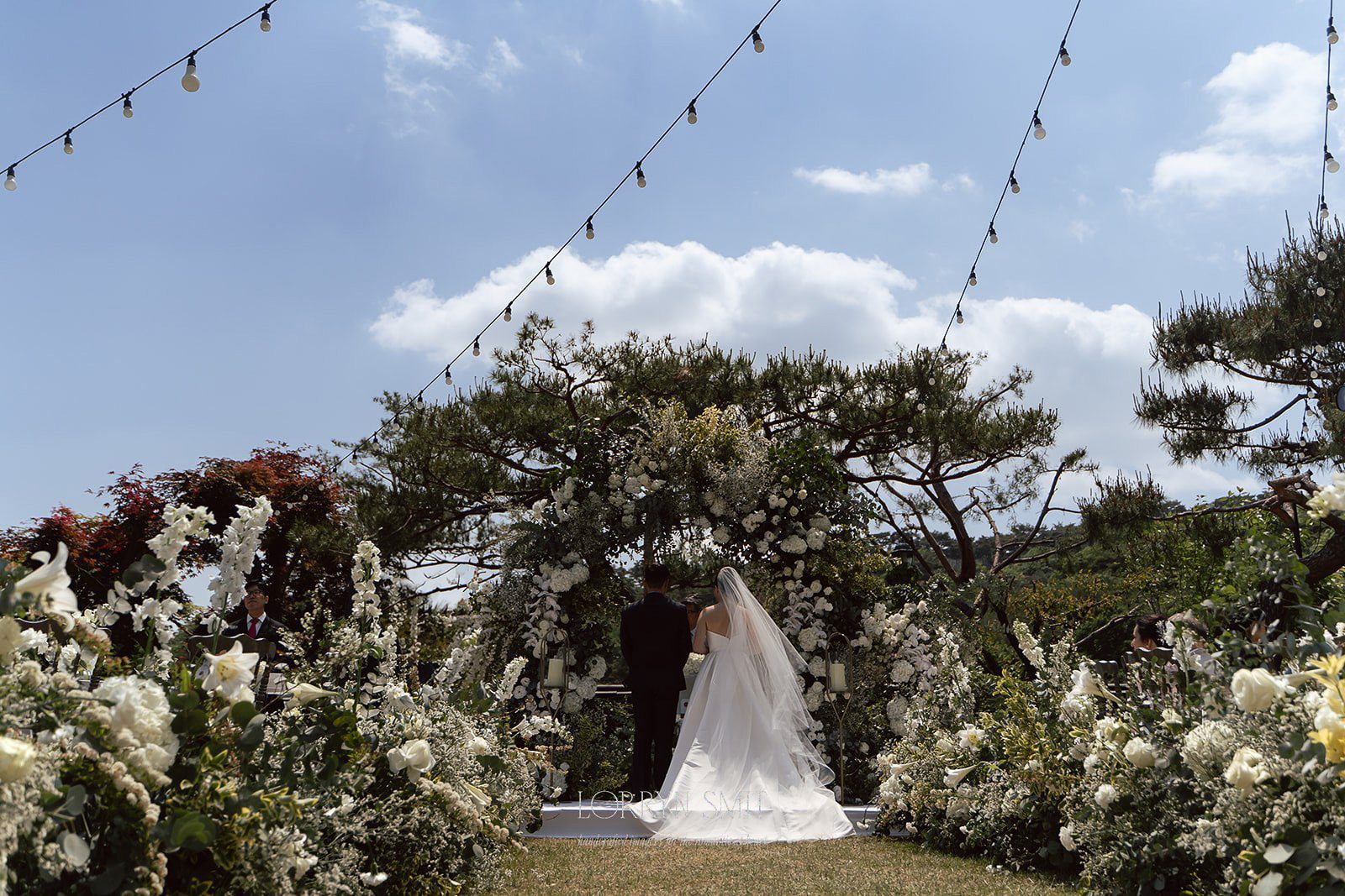 A bride and groom stand together at an outdoor wedding altar decorated with white flowers, surrounded by greenery and string lights under a blue sky—an idyllic scene often found at top wedding venues in Korea.