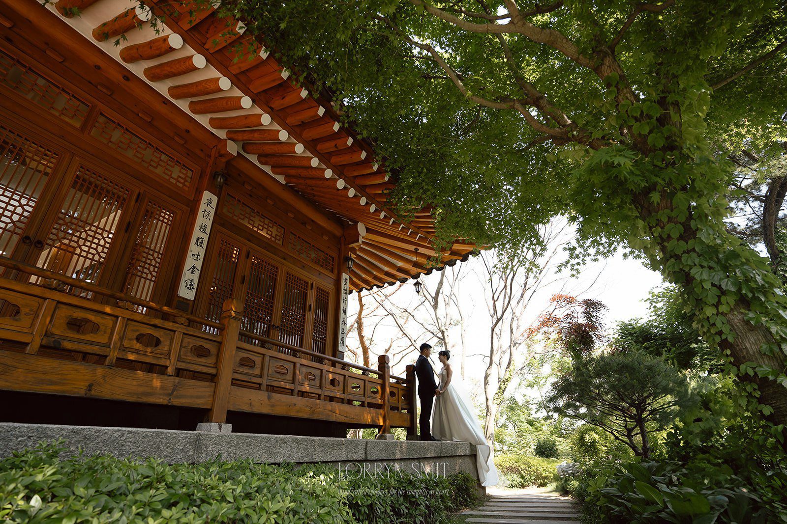 A couple in wedding attire stands outside a traditional wooden building surrounded by lush greenery and large trees, capturing the charm of wedding venues in Korea.