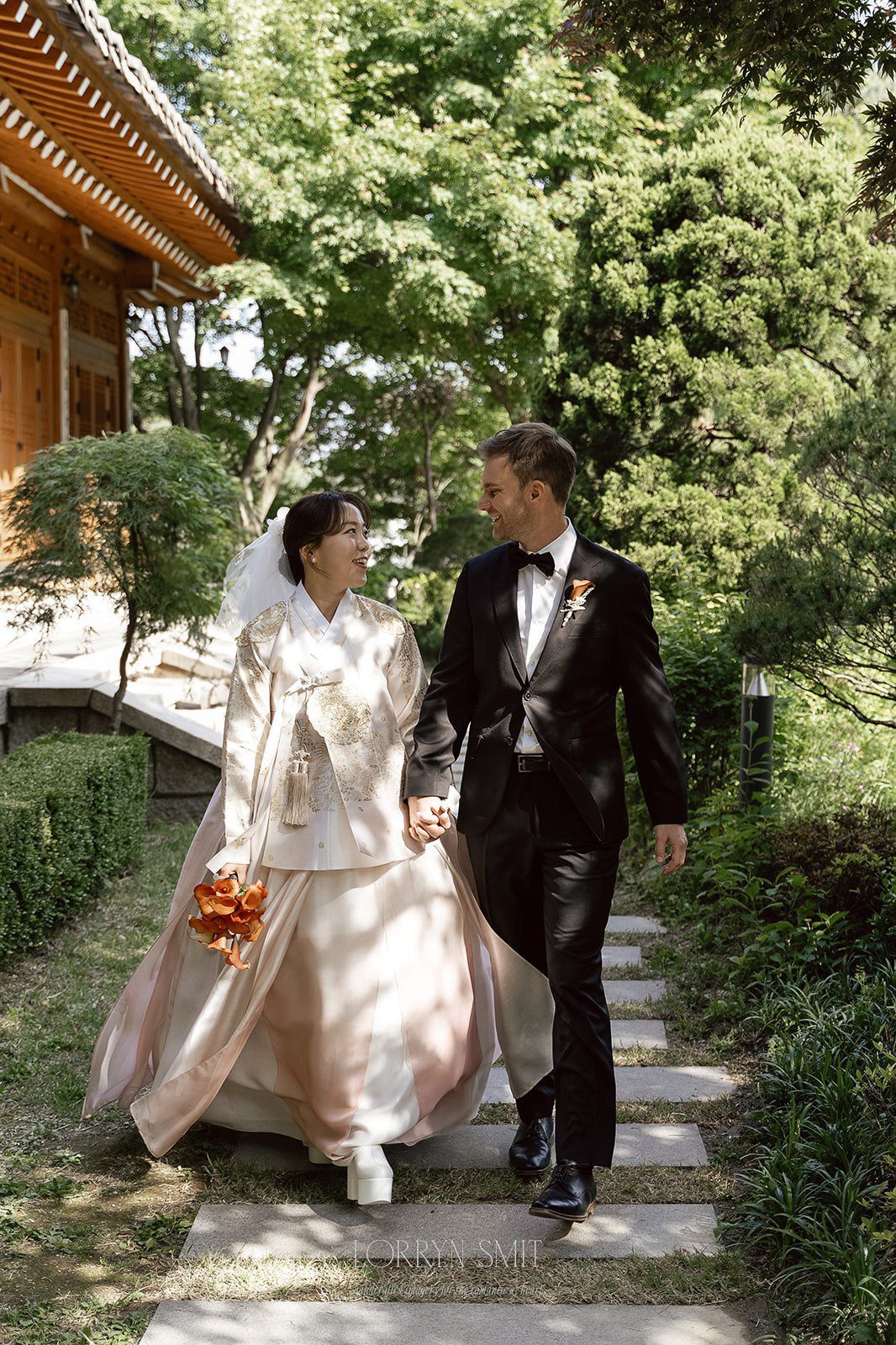A bride in a traditional Korean hanbok and a groom in a black suit walk hand in hand along a stone path in a garden, showcasing the elegance of wedding venues in Korea.