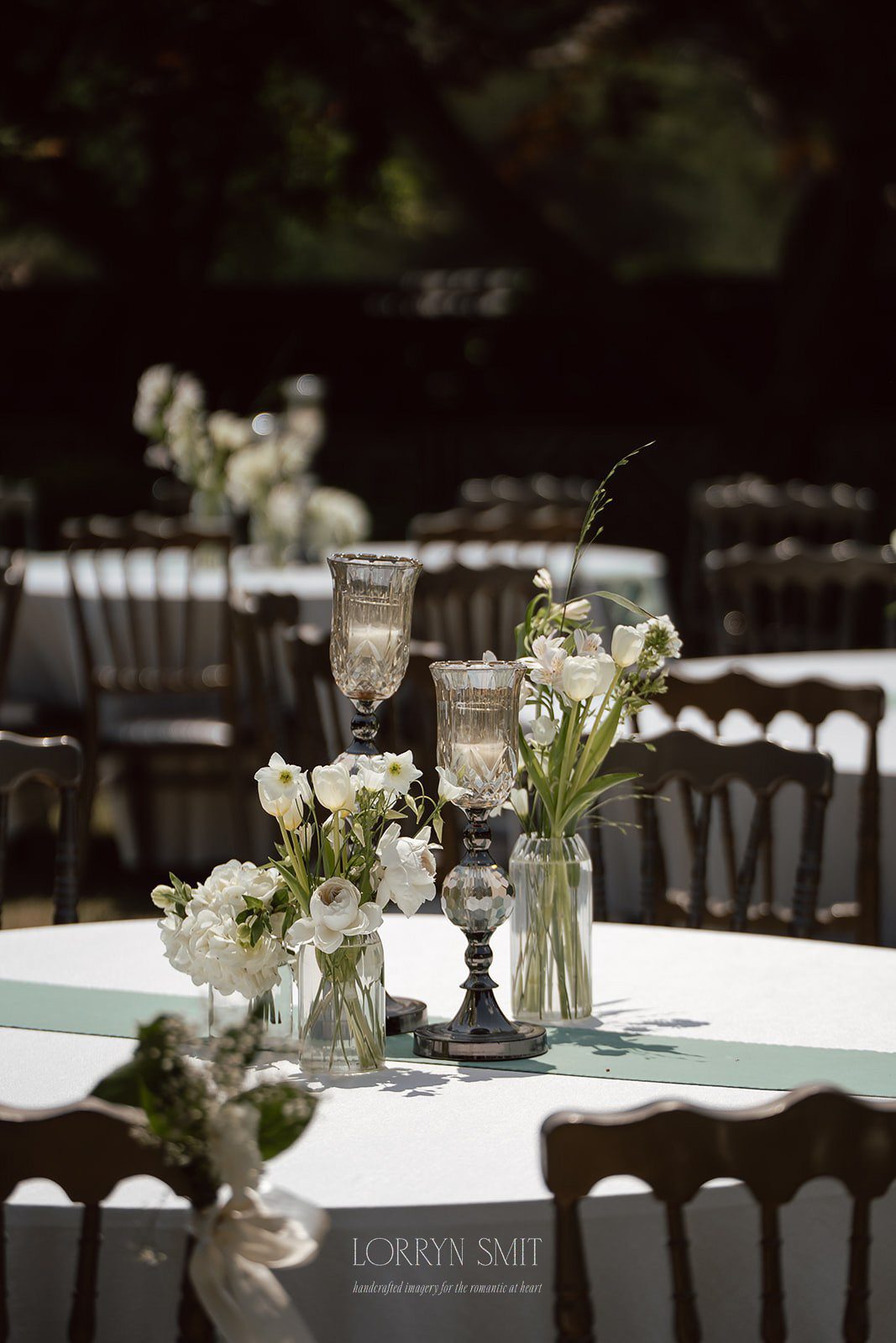Outdoor event table with white tablecloth, vintage glass candle holders, and clear vases of white flowers.