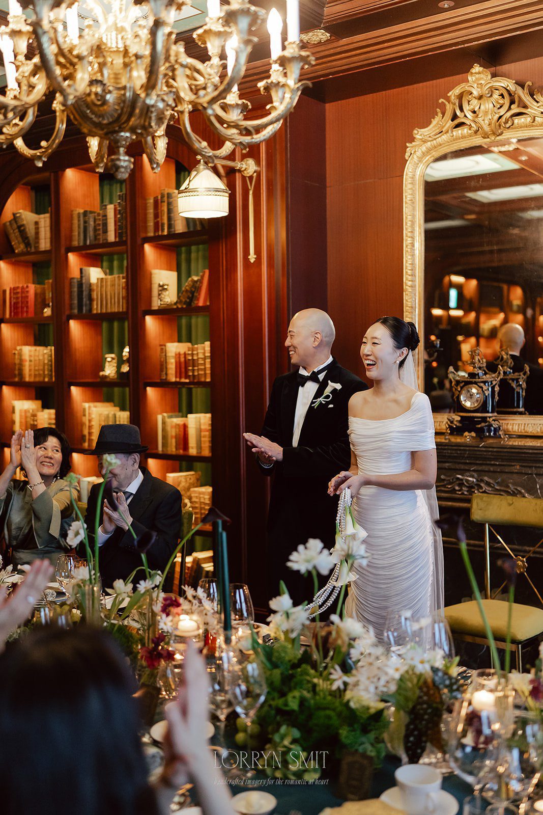 A bride and groom stand smiling in an elegant, wood-paneled room—one of the stunning wedding venues in Korea—as seated guests applaud. The table is set with flowers and glassware, and bookshelves line the background.