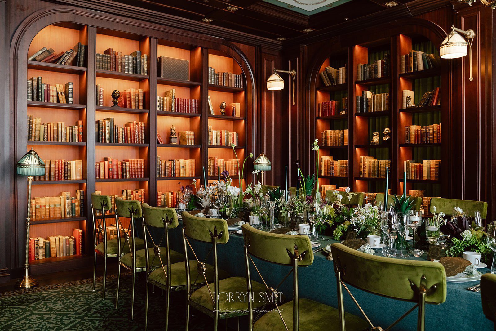 A long dining table with green velvet chairs is elegantly set for a meal in a library room.