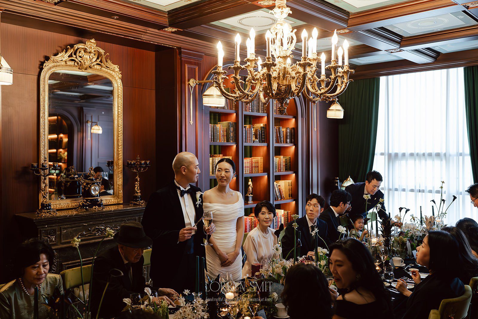 A bride and groom stand and smile at a formal indoor reception, surrounded by seated guests in an elegant, wood-paneled room with chandeliers and bookshelves—one of the most enchanting wedding venues in Korea.