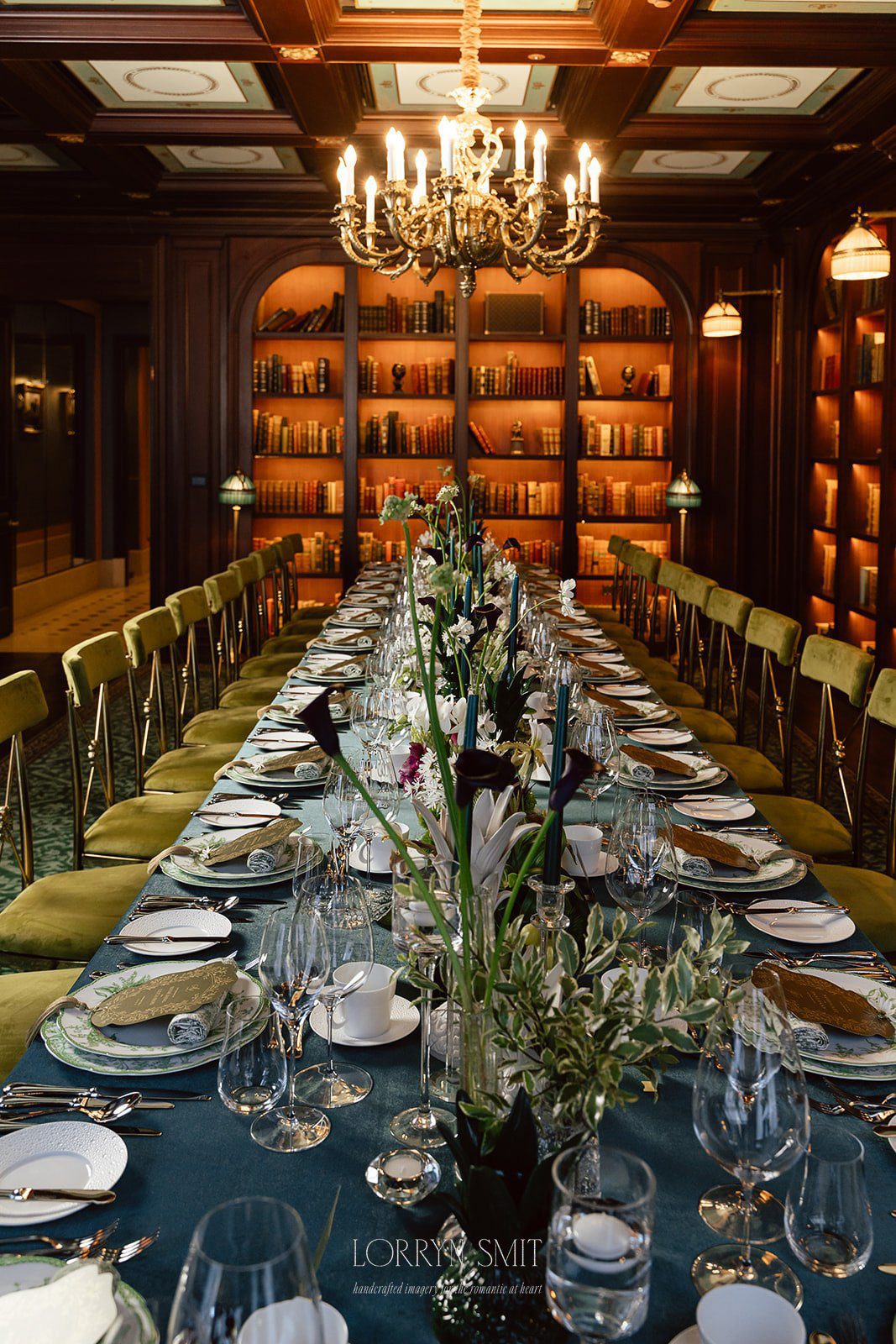 A long dining table is elegantly set with plates, glasses, and flowers in a room with bookshelves, wood paneling, green chairs, and a chandelier—an atmosphere reminiscent of top wedding venues in Korea.