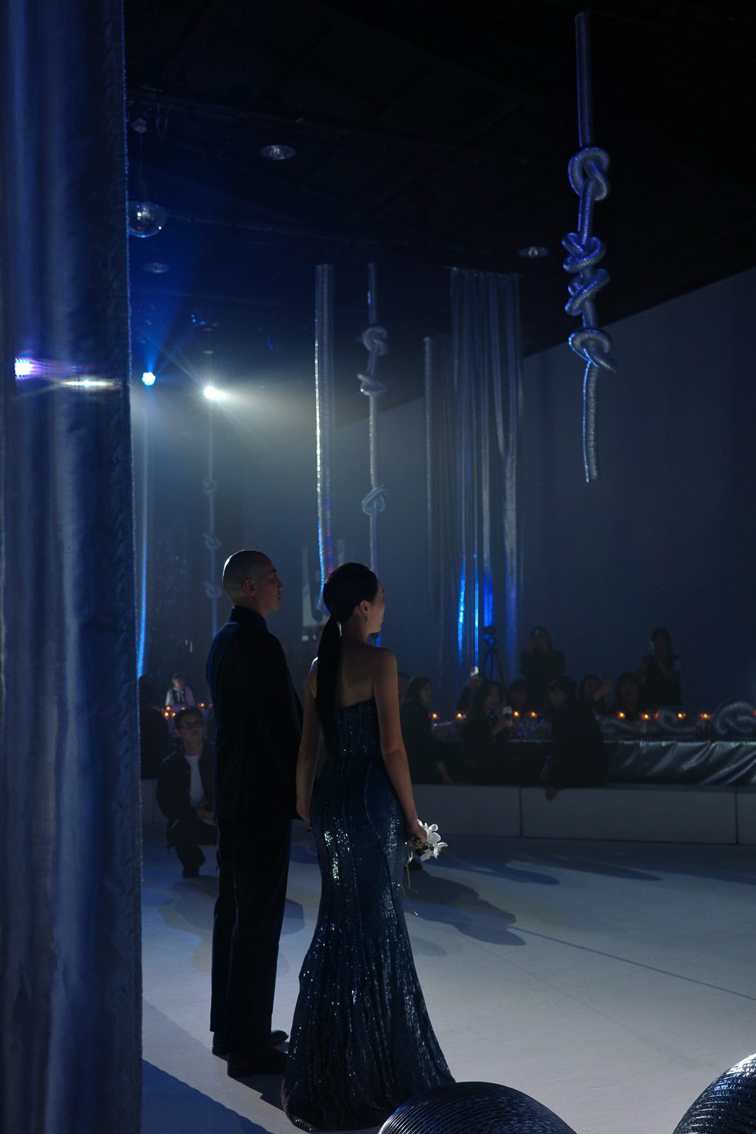 A man and woman in formal evening attire stand on a dimly lit stage with hanging rope decorations, facing an audience seated at candlelit tables—captured perfectly by what is a wedding content creator.