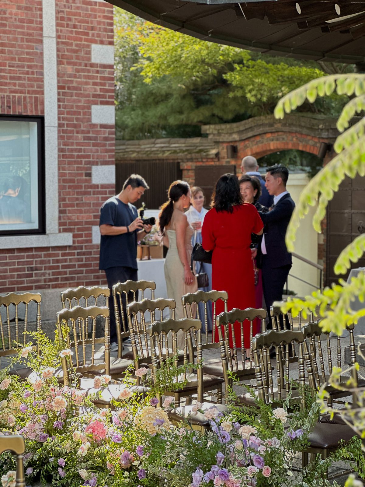 A group of people converse outdoors near gold chairs and a floral arrangement, with a brick building and trees in the background.