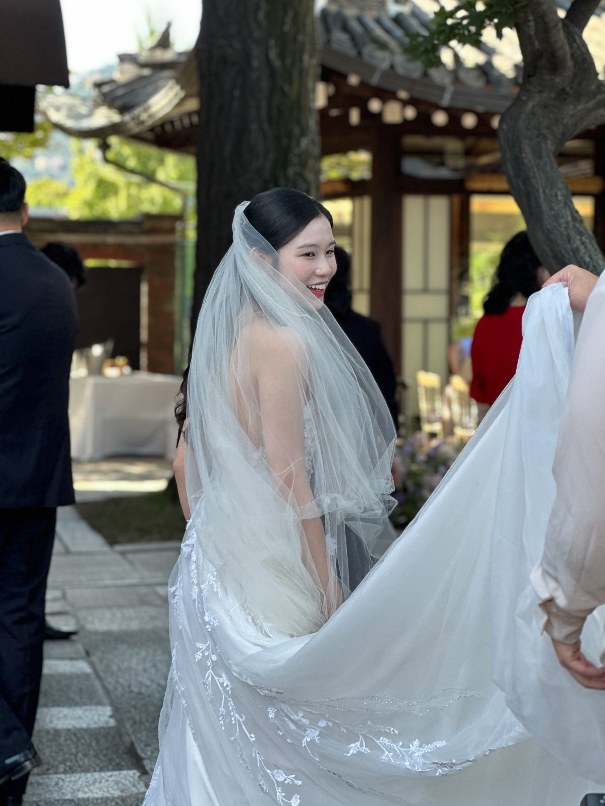 A bride in a white wedding dress and veil smiles as someone holds her train during an outdoor ceremony near a traditional building, capturing moments perfect for anyone wondering what is a wedding content creator.