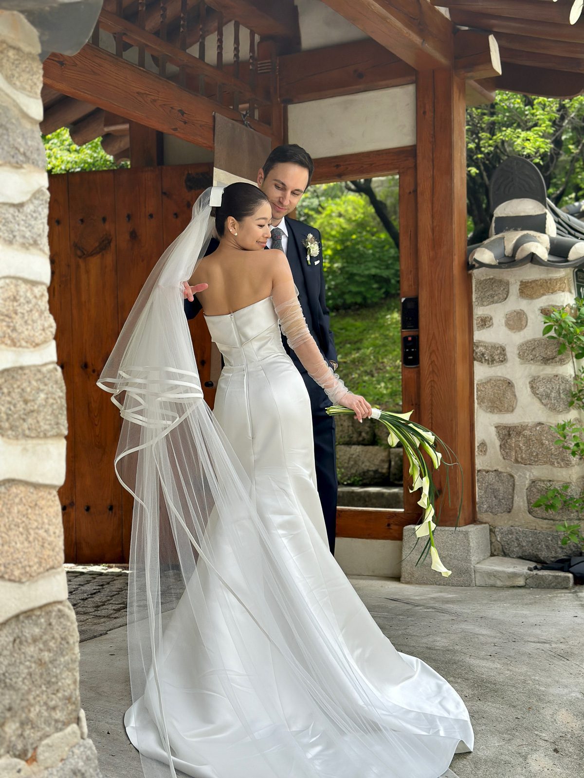 A bride in a white gown and veil holds flowers while standing beside a groom in a suit under a wooden and stone structure outdoors, capturing precious memories—perhaps with the help of a wedding content creator.