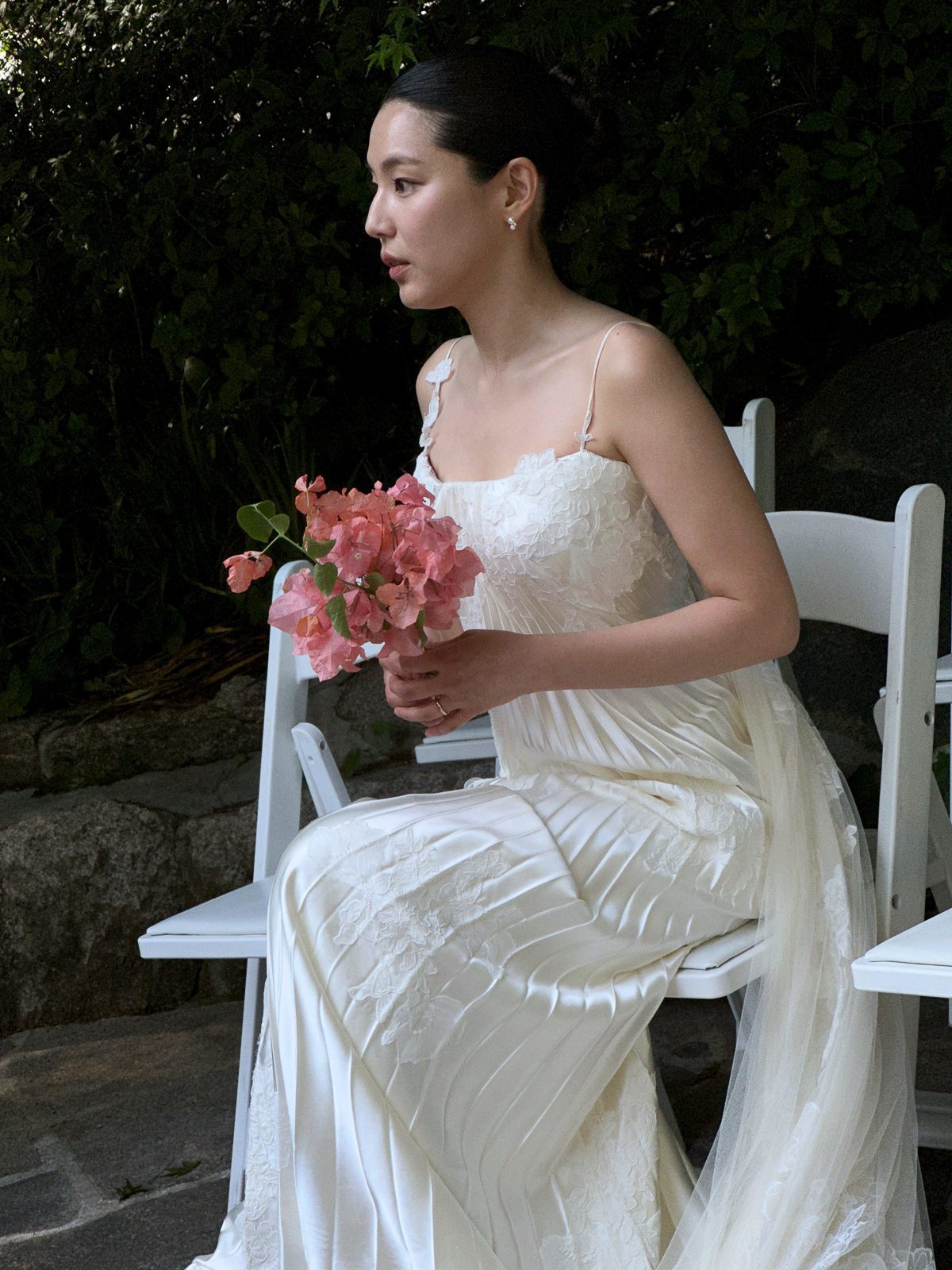 A woman in a white dress holds a bouquet of pink flowers while seated on a white chair outdoors, capturing the elegance that inspires anyone wondering what is a wedding content creator.
